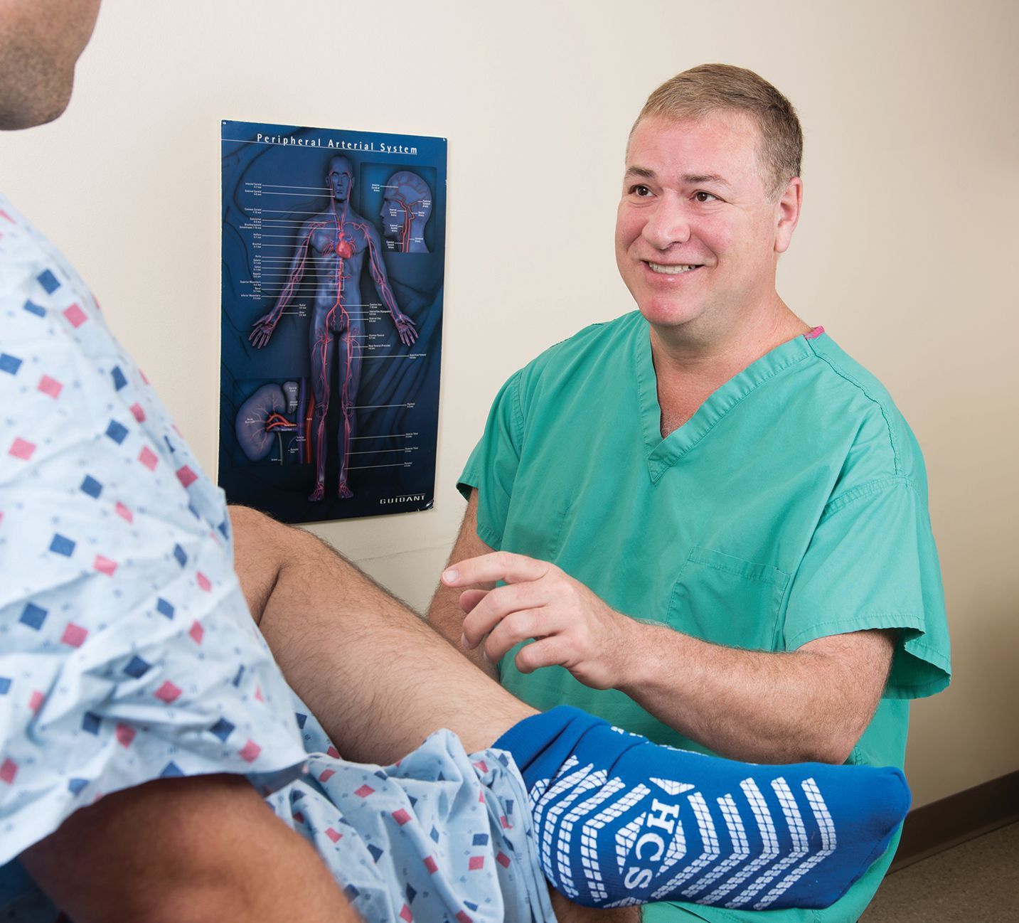 A medical professional in green scrubs talks to a patient wearing a hospital gown and blue non-slip socks in an exam room.