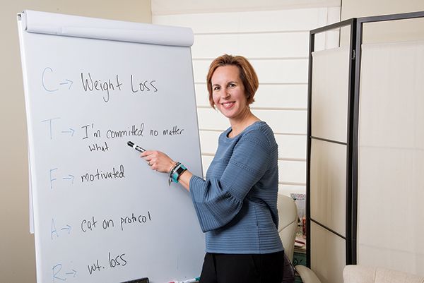 A person in a blue top stands by a whiteboard, pointing to a list of weight loss commitments written on the board.