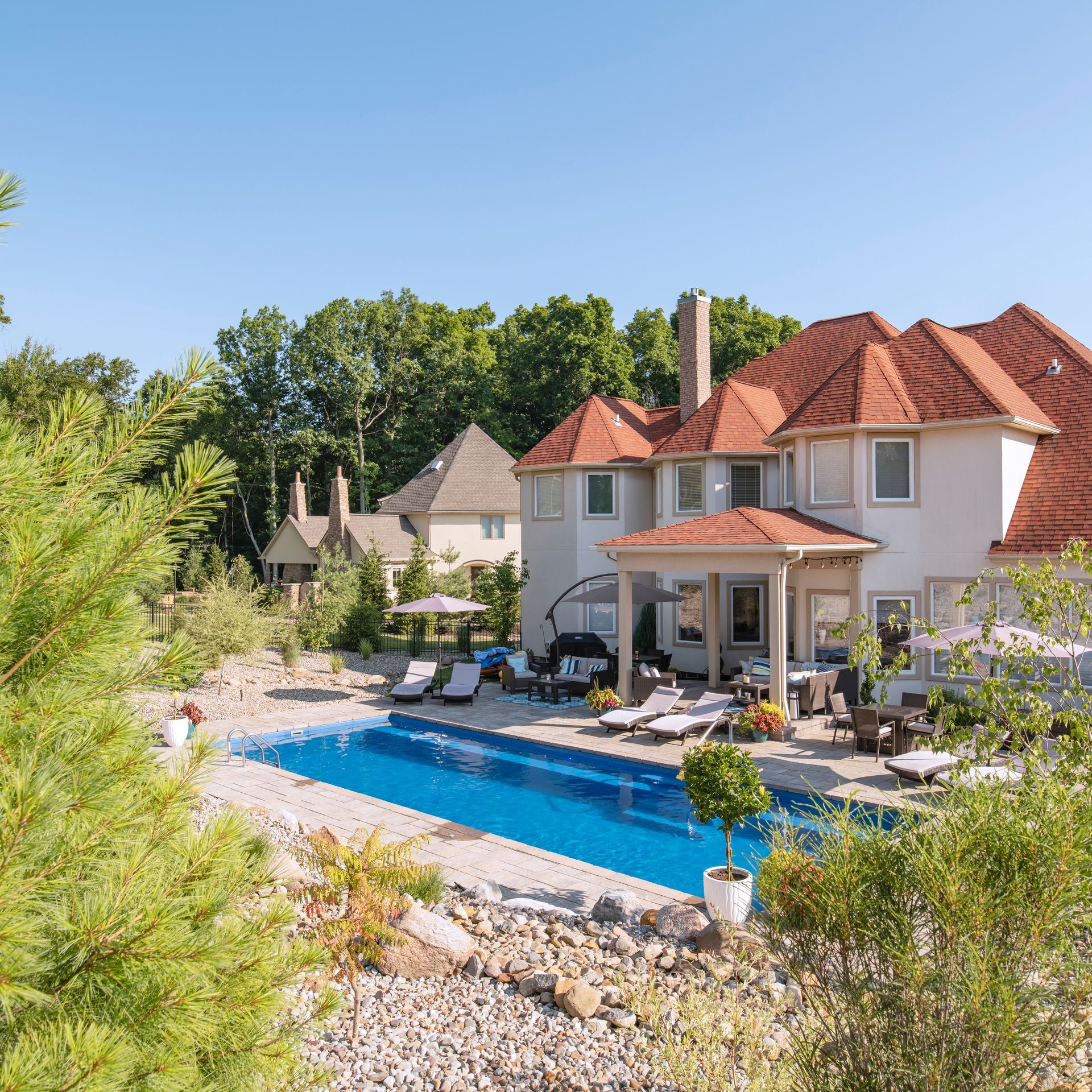 A large, multi-story house with a terracotta roof sits beside a rectangular blue swimming pool on a sunny day.