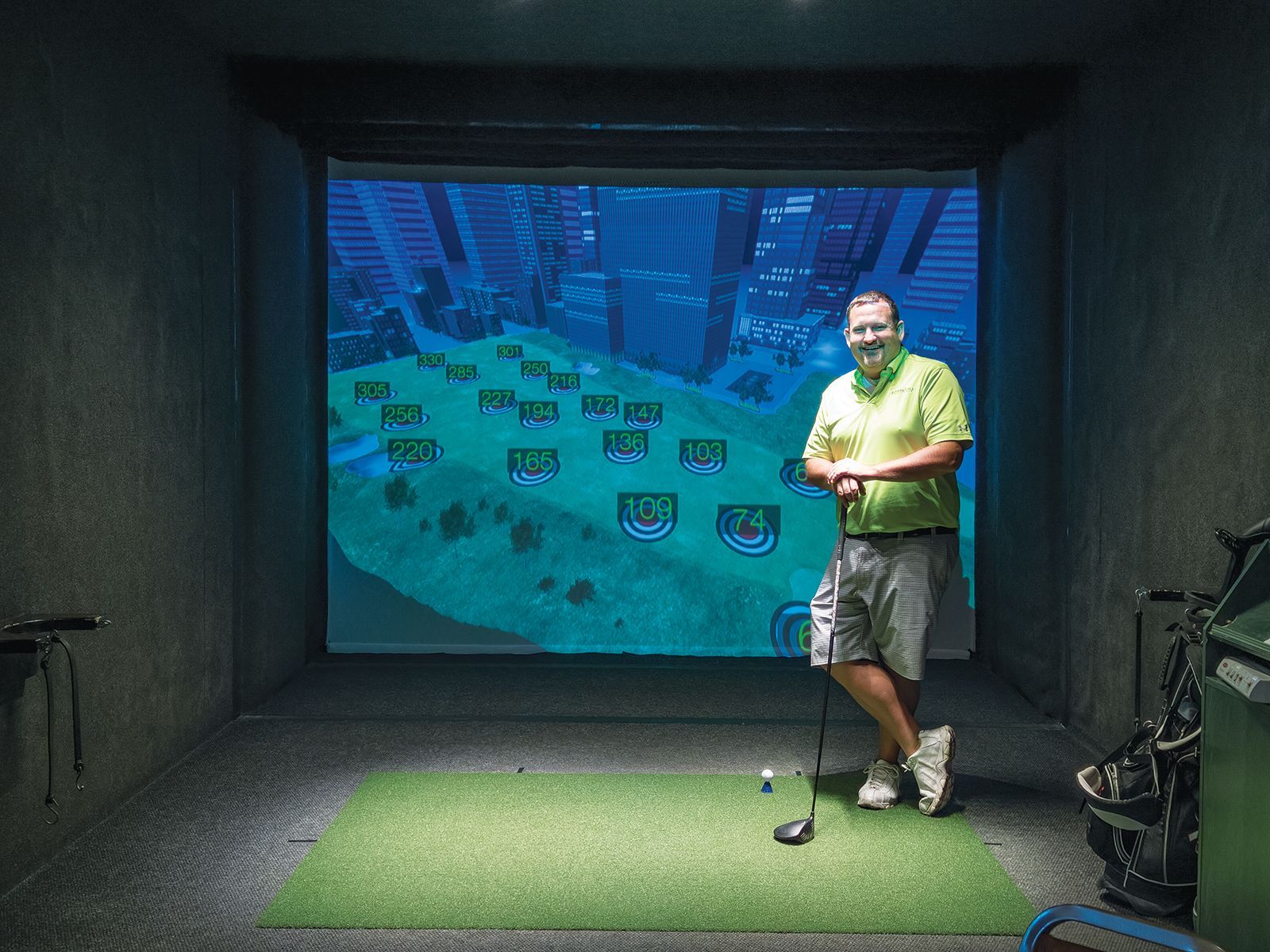 A smiling person in a bright green shirt stands on a putting mat in front of a golf simulator screen displaying targets.