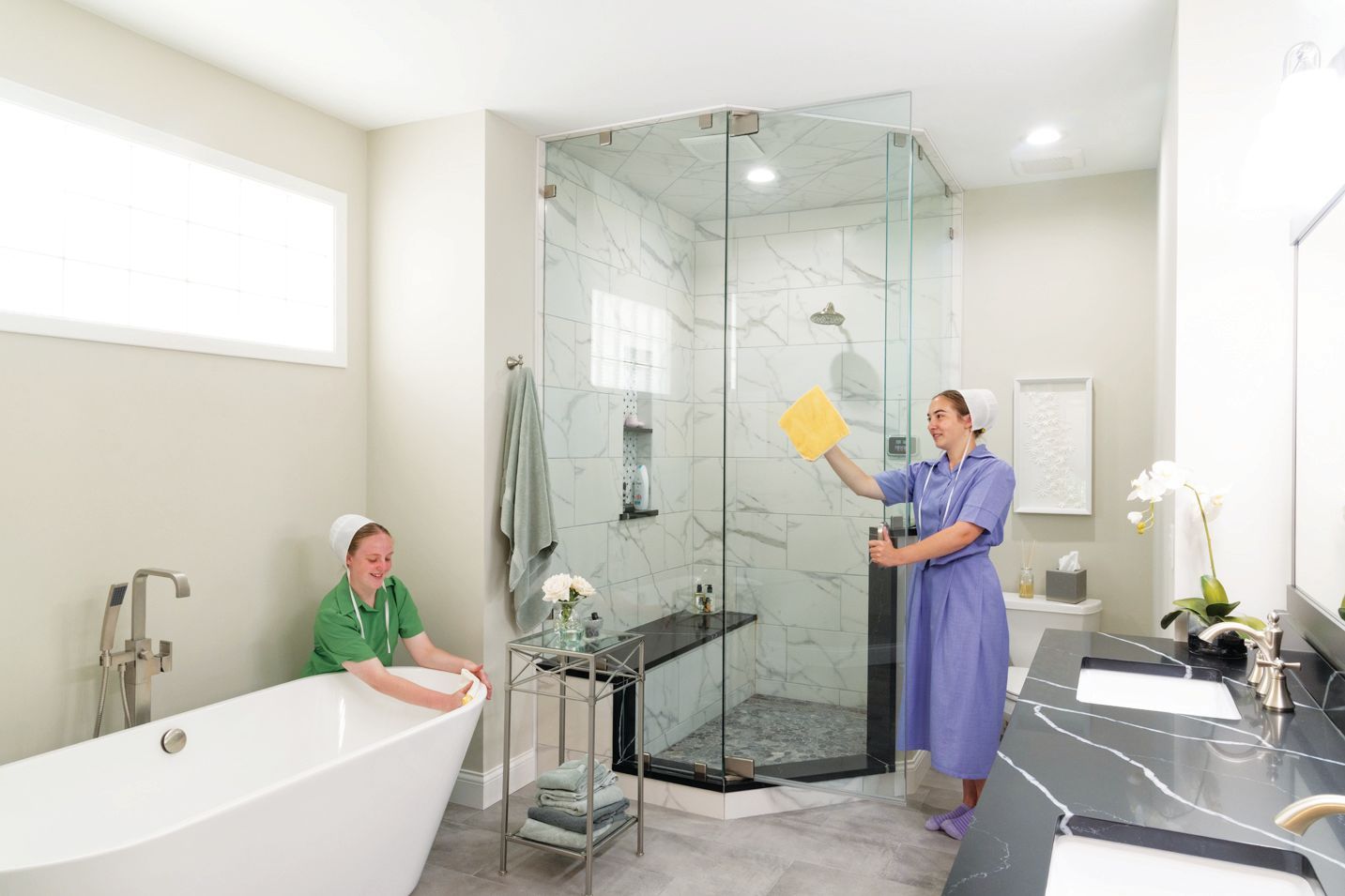 Two professionals in uniforms clean a modern bathroom, one wiping the glass shower and the other inspecting the bathtub.