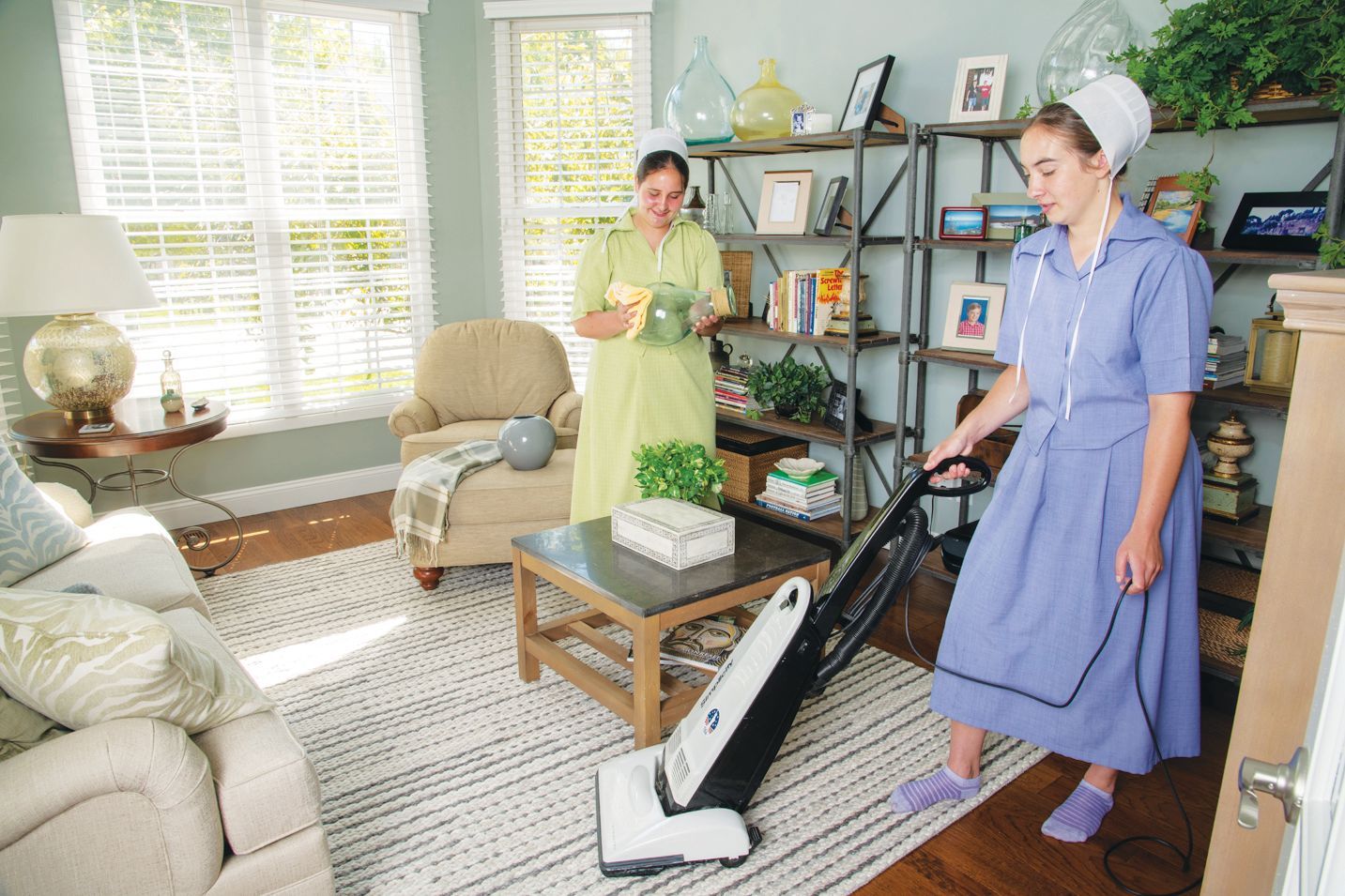 Two individuals in modest dresses and caps clean a bright, furnished living room: one vacuums while the other dusts a shelf.