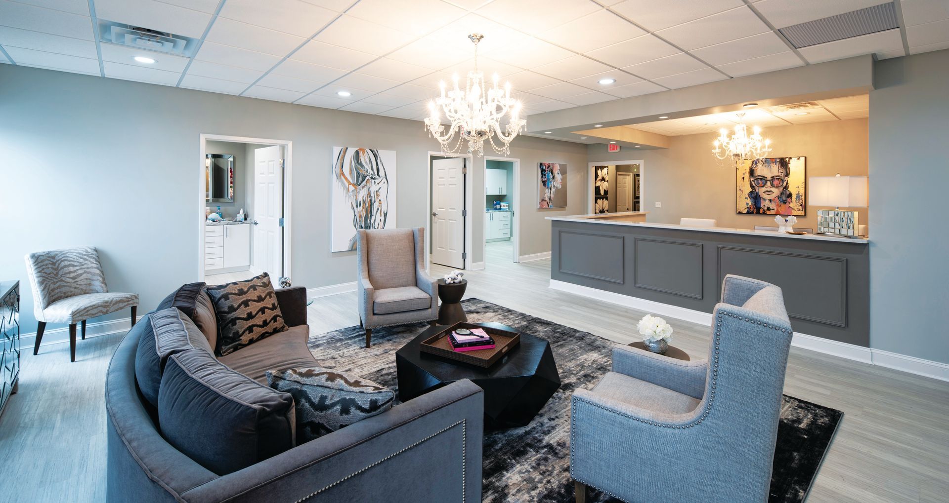 A modern, elegant waiting room with a reception desk, gray armchair seating, a central coffee table, and chandeliers.