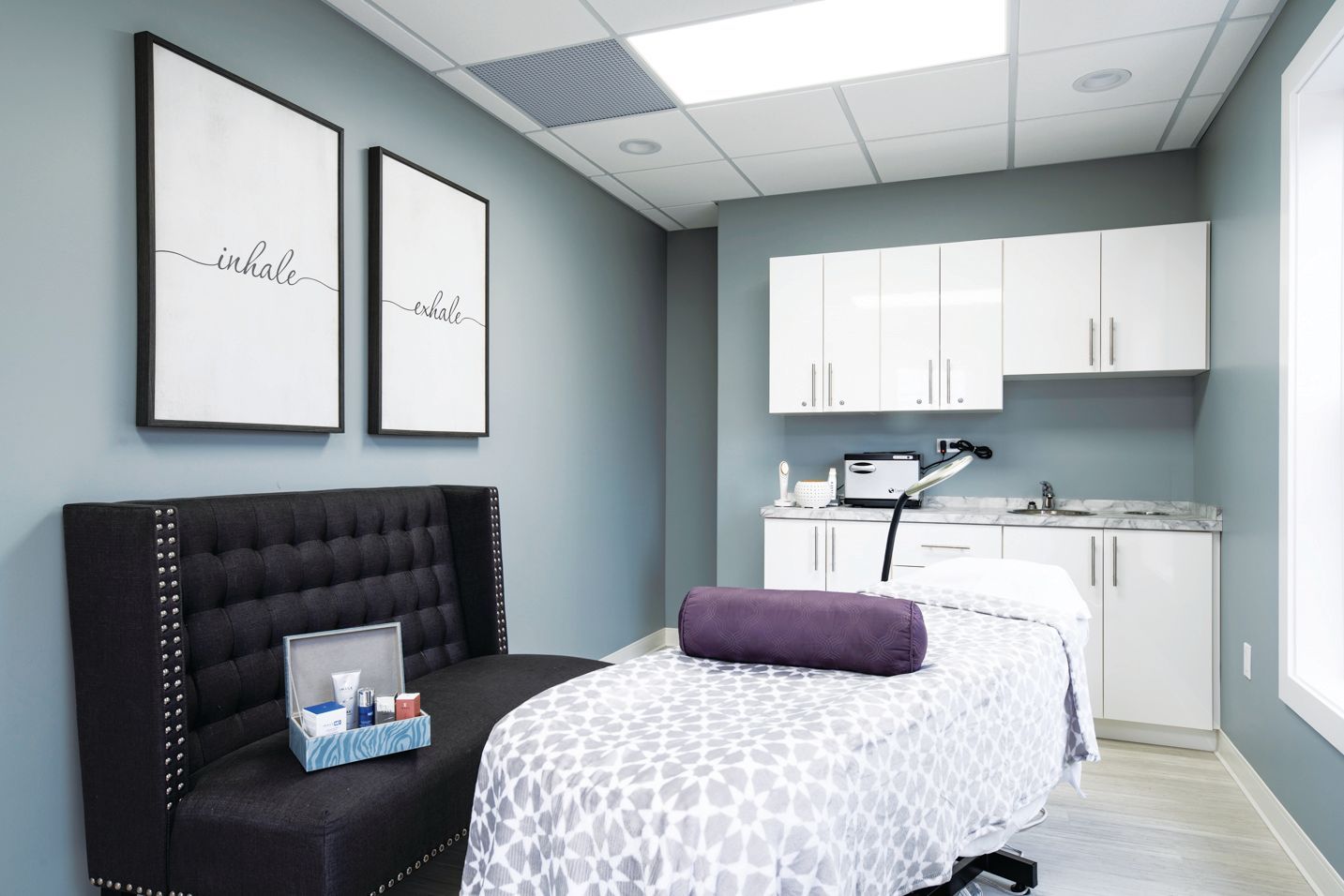 Modern med spa treatment room with a massage bed, tufted bench, white cabinets, and blue-gray walls.