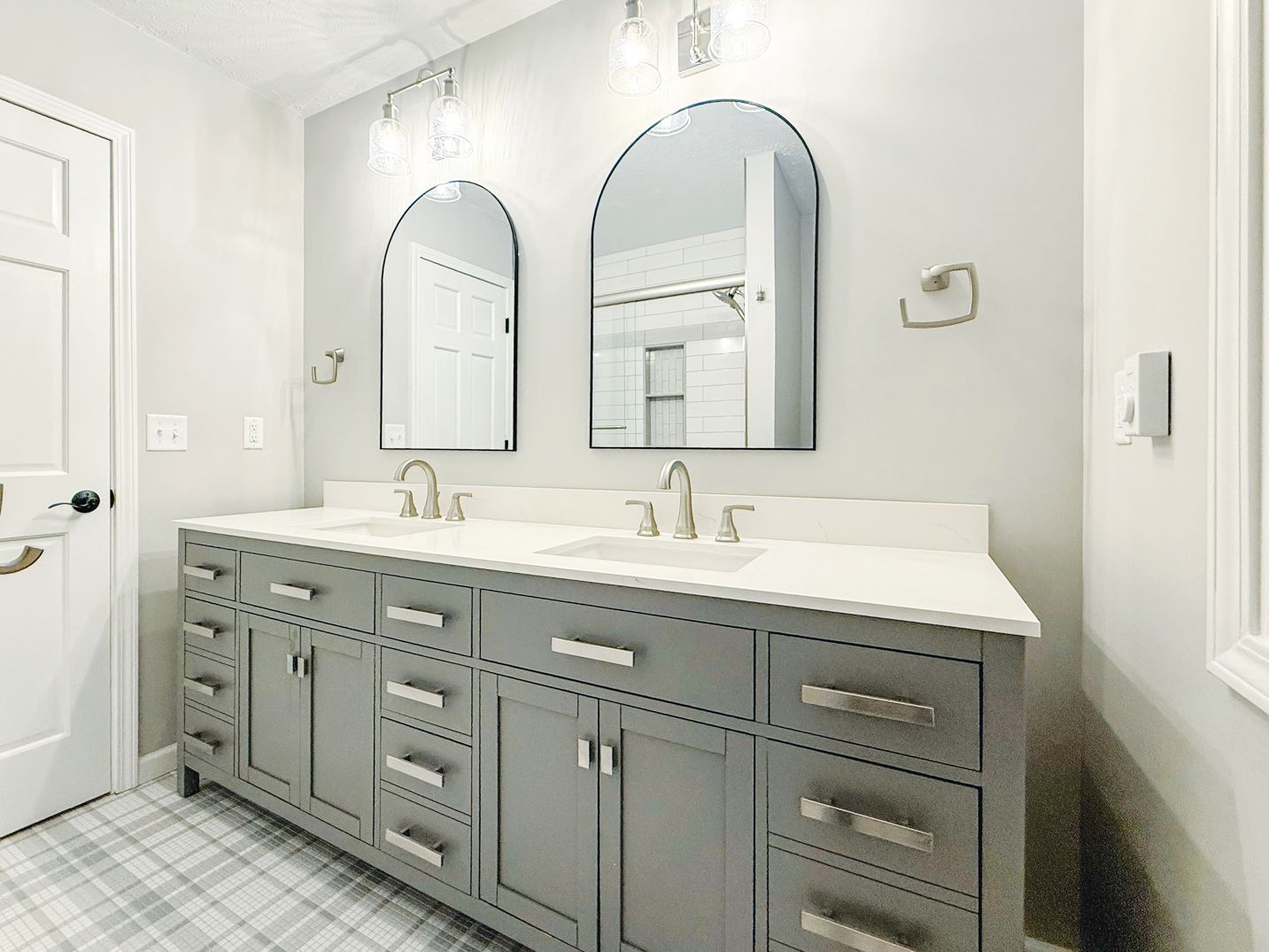 A double vanity with grey cabinets, white countertops, two arched mirrors, and matching wall sconces in a bright bathroom.