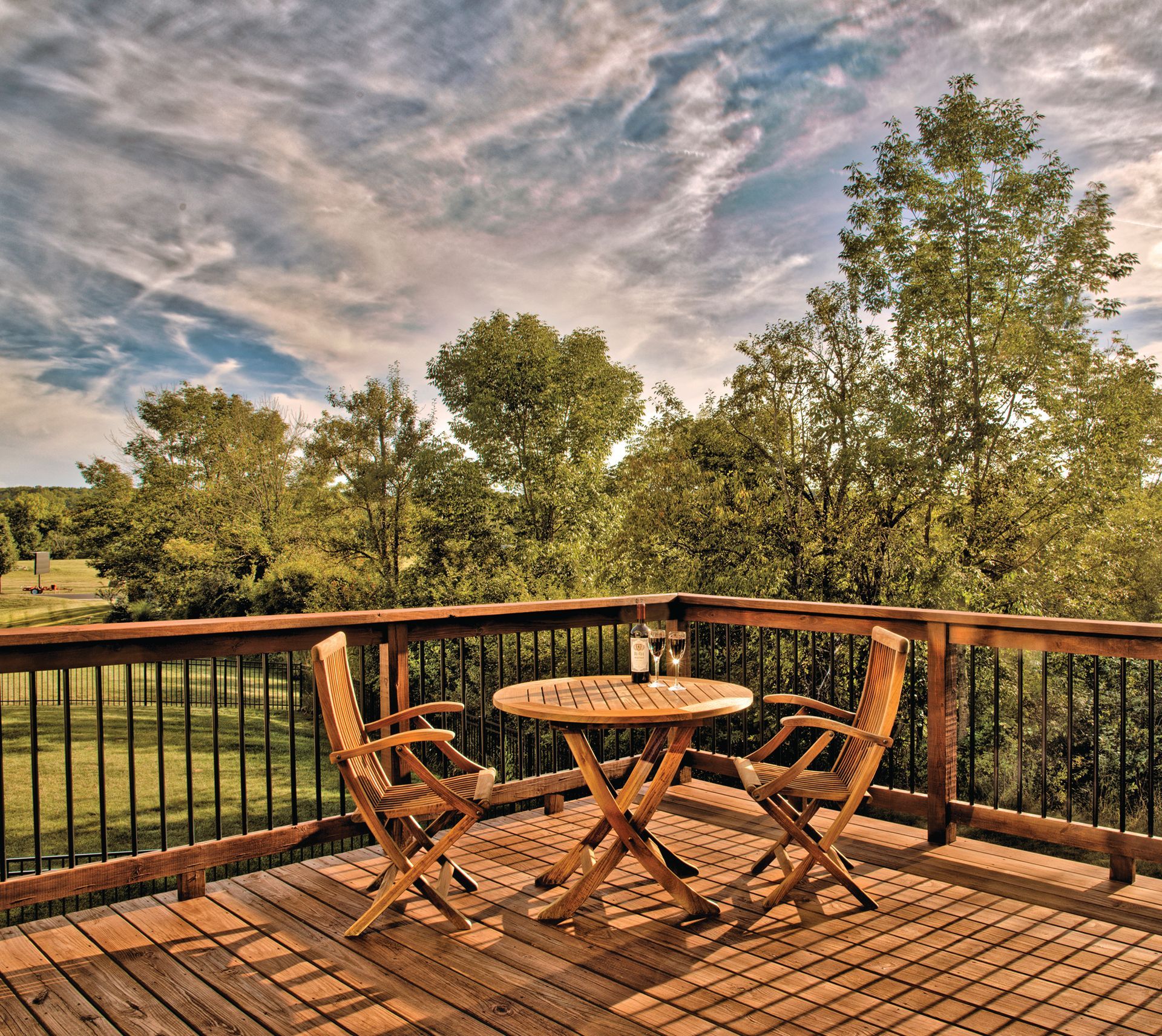 A wooden deck with a small round table and two folding chairs, overlooking trees and a grassy field under a cloudy sky.