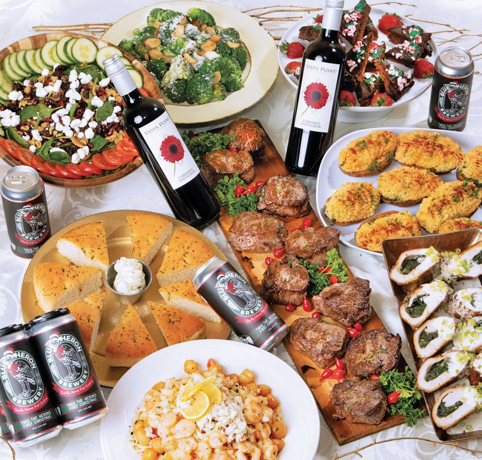 A top-down view of a lavish dinner spread featuring roasted meats, pasta, salad, flatbread, wine, and craft beer cans.