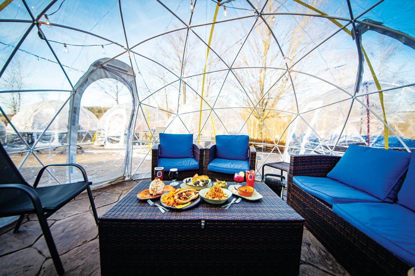 A clear dome structure containing a patio table with food, blue cushioned chairs, and a wicker sofa on a stone floor.