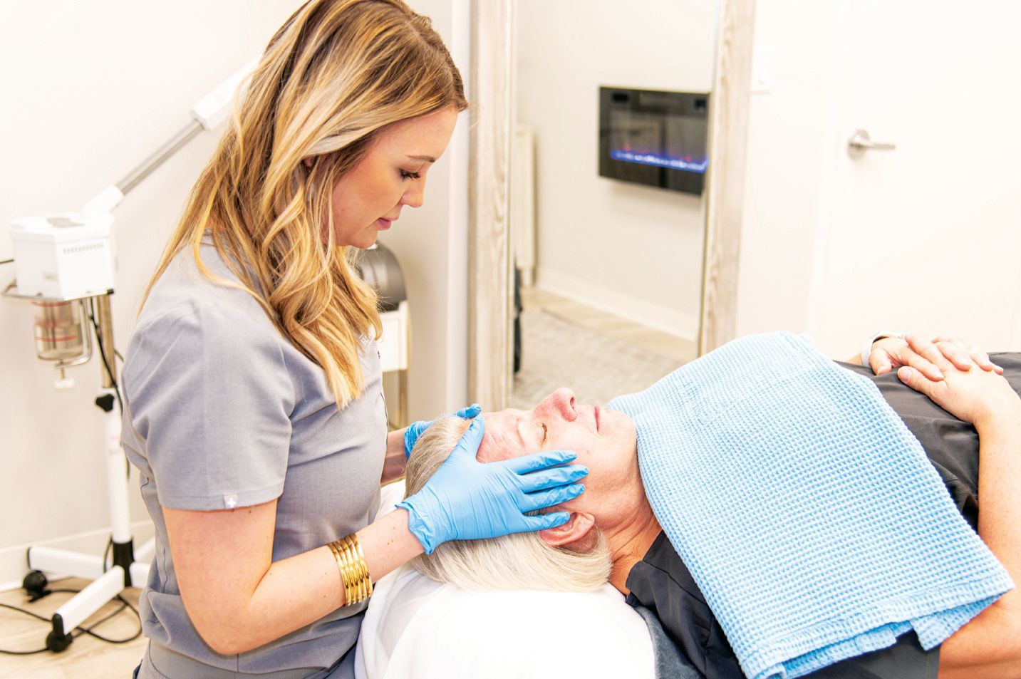 A technician in blue gloves performs a facial treatment on a client lying on a treatment table.