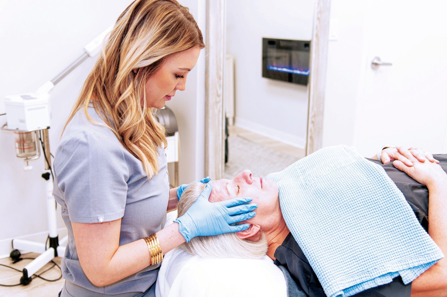 A person wearing blue gloves performs a facial treatment on a client lying on a spa bed in a bright, modern room.