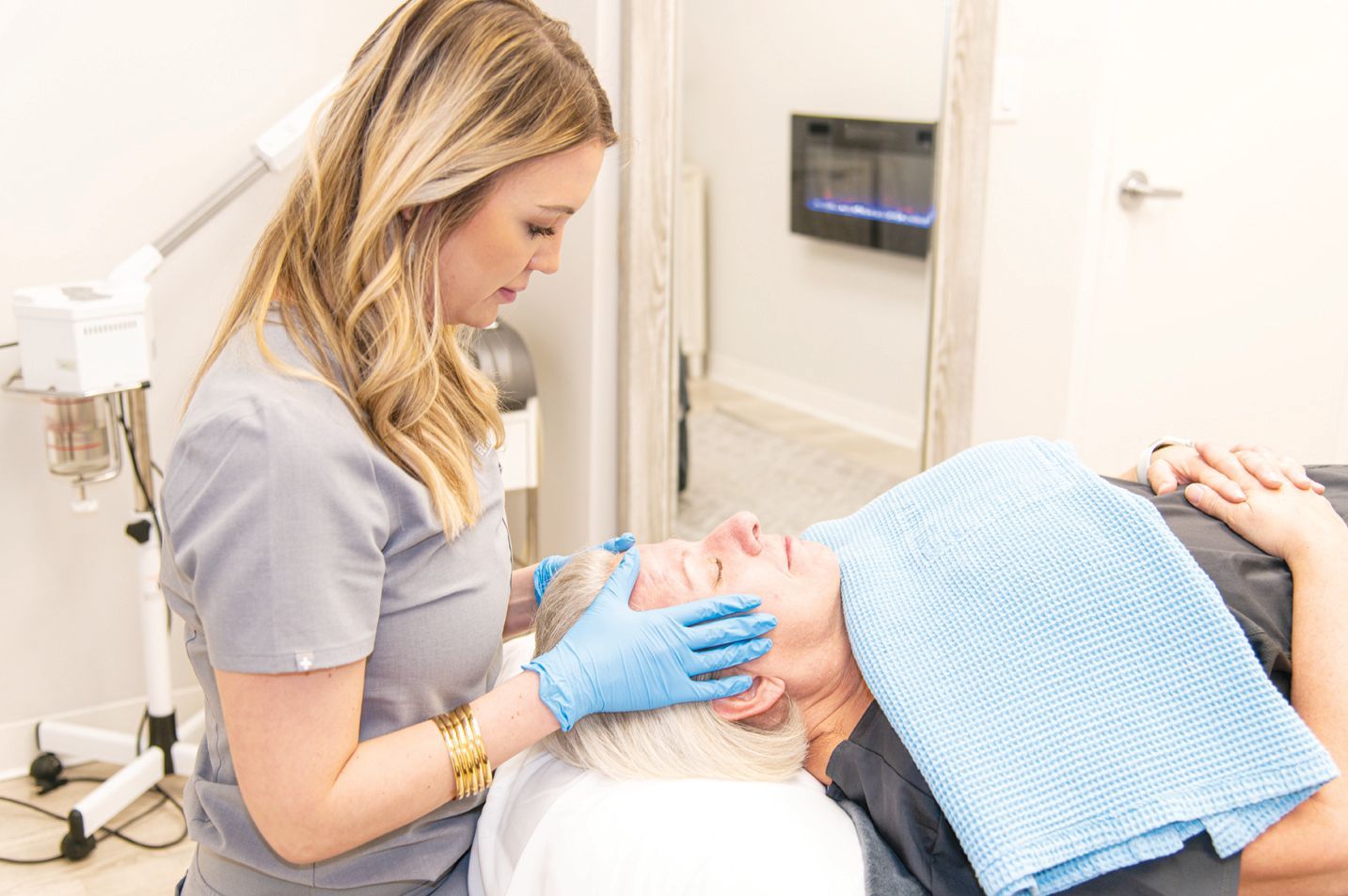 An aesthetician wearing blue gloves provides a facial massage to a client lying on a treatment table.