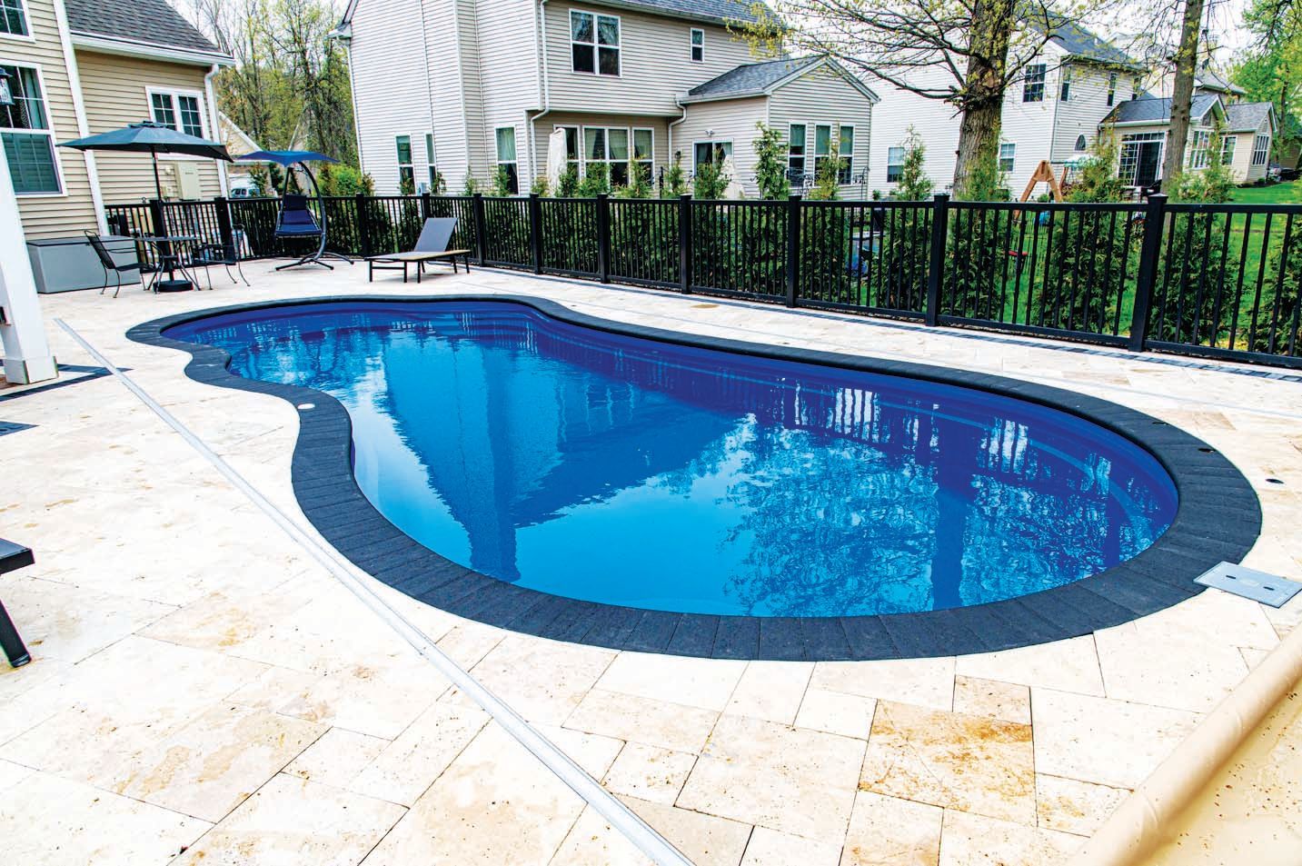 A kidney-shaped swimming pool with blue water surrounded by light-colored stone pavers and a black metal fence.