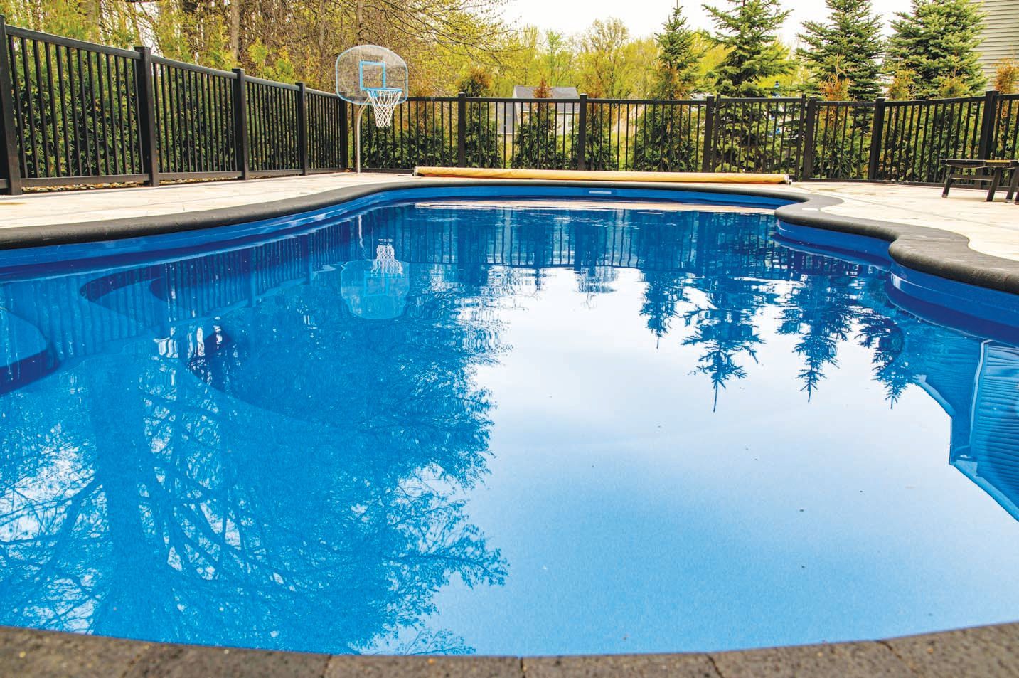 A clear, bright blue swimming pool with a dark stone border, surrounded by a black fence and evergreen trees.