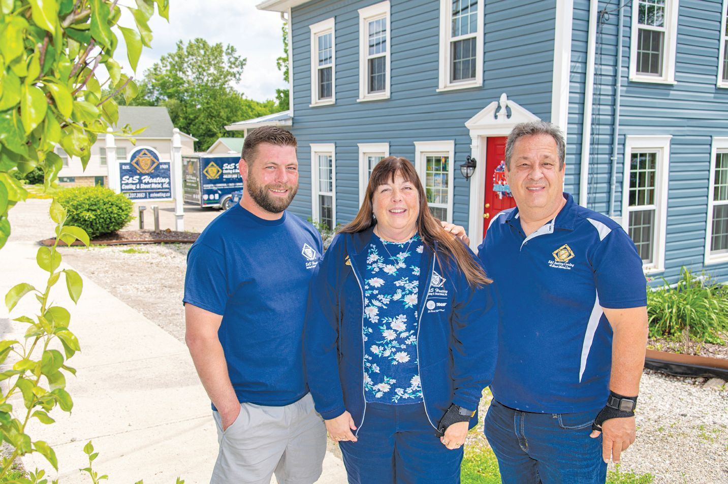 Three people in blue uniforms stand in front of a blue house with a red door, smiling for a photo.