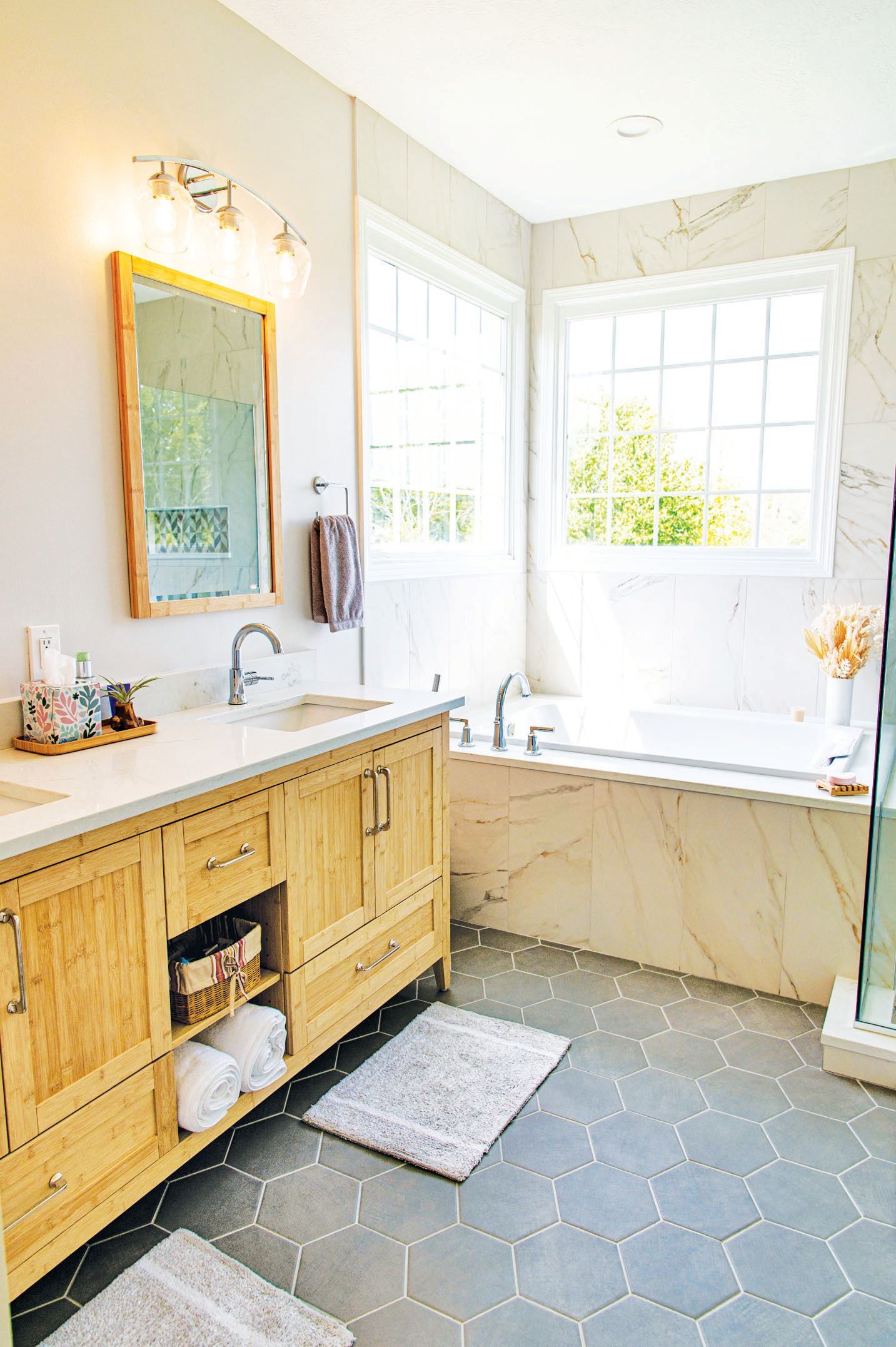 A bright bathroom with a light wood vanity, grey hexagonal floor tiles, and a large bathtub by a window.