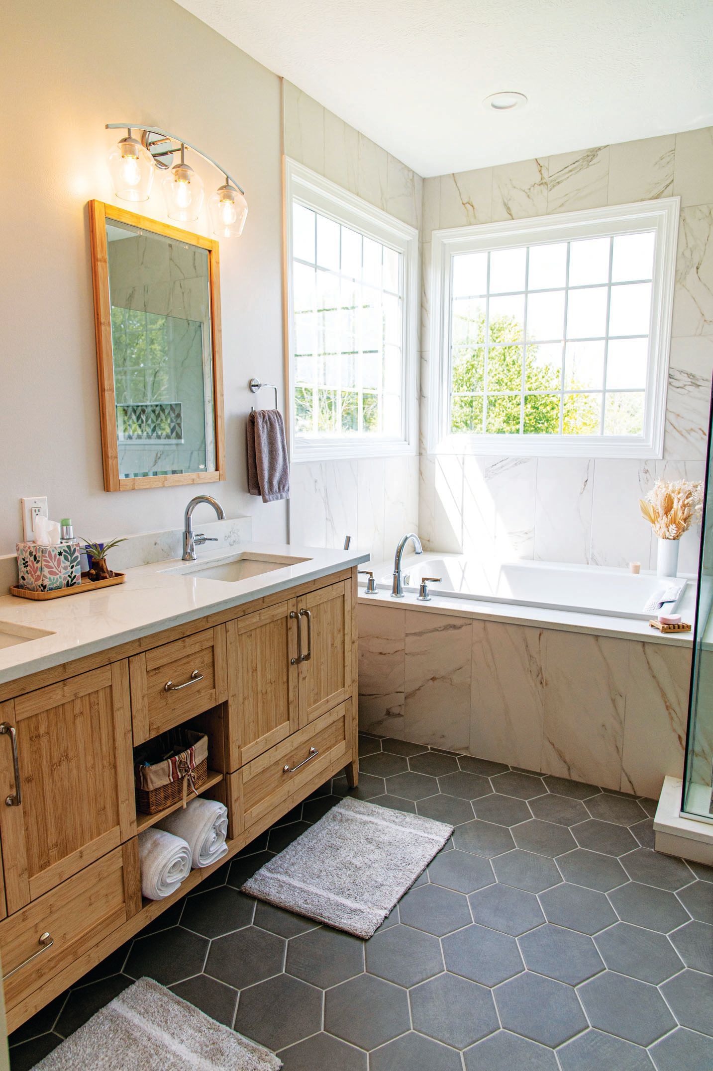 Modern bathroom with wooden vanity, marble-tiled tub, large windows, and grey hexagonal floor tiles.