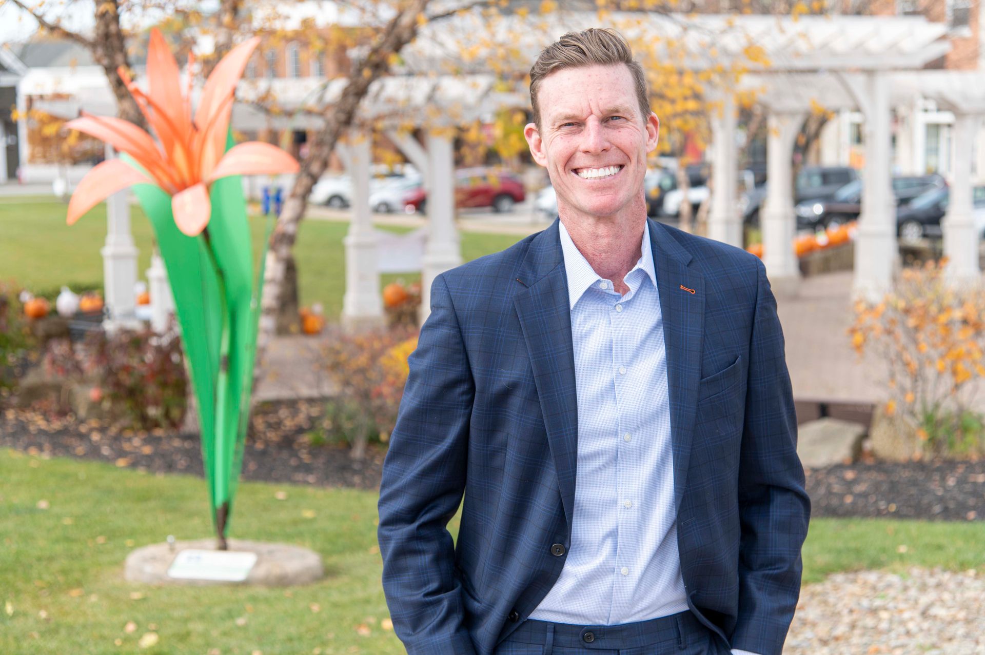 A smiling person in a blue suit and light shirt stands outside near a large orange metal flower sculpture.