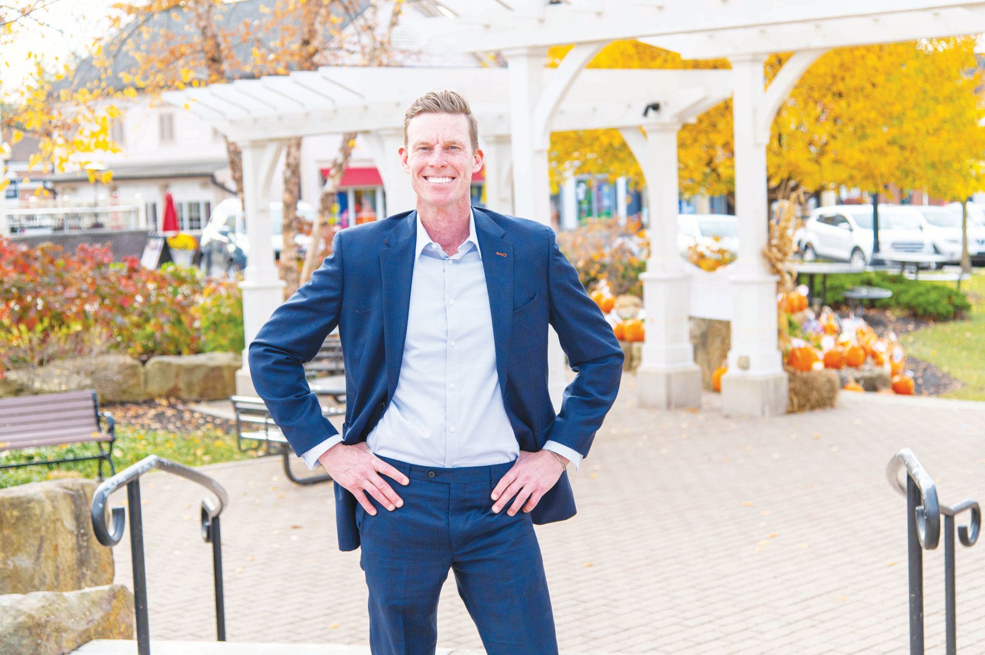 A smiling person in a blue suit stands with hands on hips under a white trellis in a park with autumn foliage.