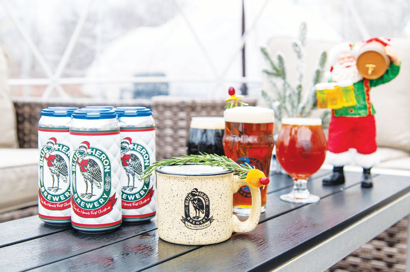 Cans of Port Huron Brewing Company beer, assorted glasses of beer, a mug, and a Santa figurine on an outdoor table.