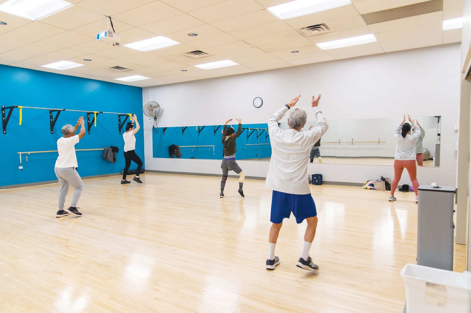 Five people participate in a fitness class in a studio with a blue wall, mirrors, and wood floors, performing exercises.