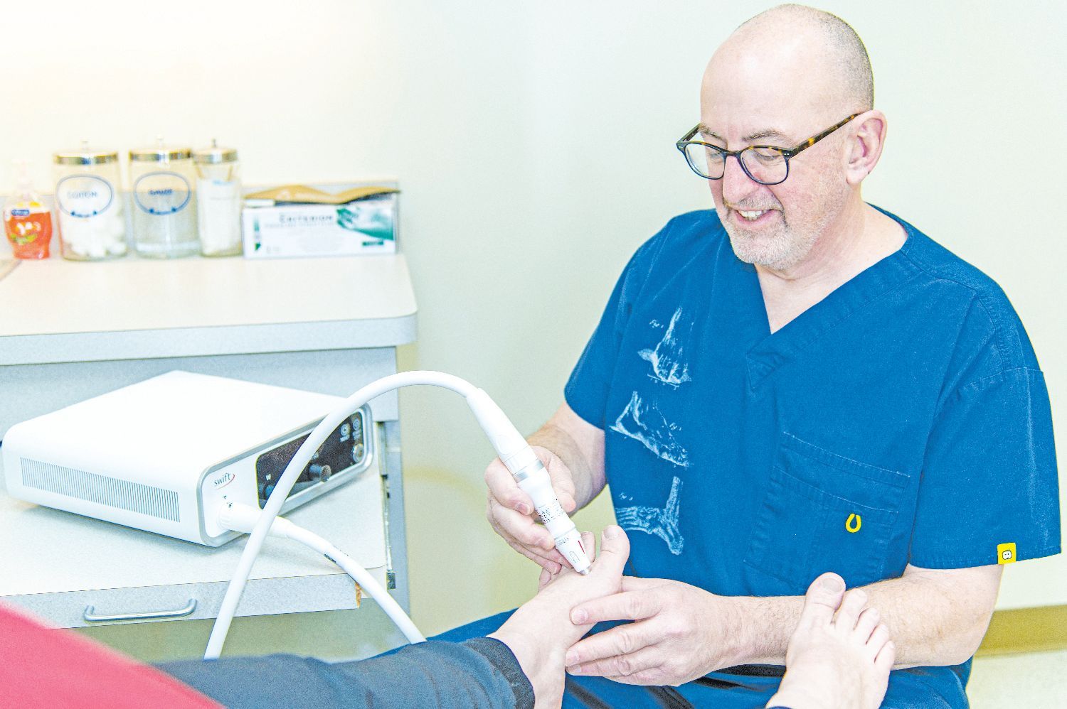 A clinician in blue scrubs uses a handheld medical device to treat a patient's foot in a clinical setting.