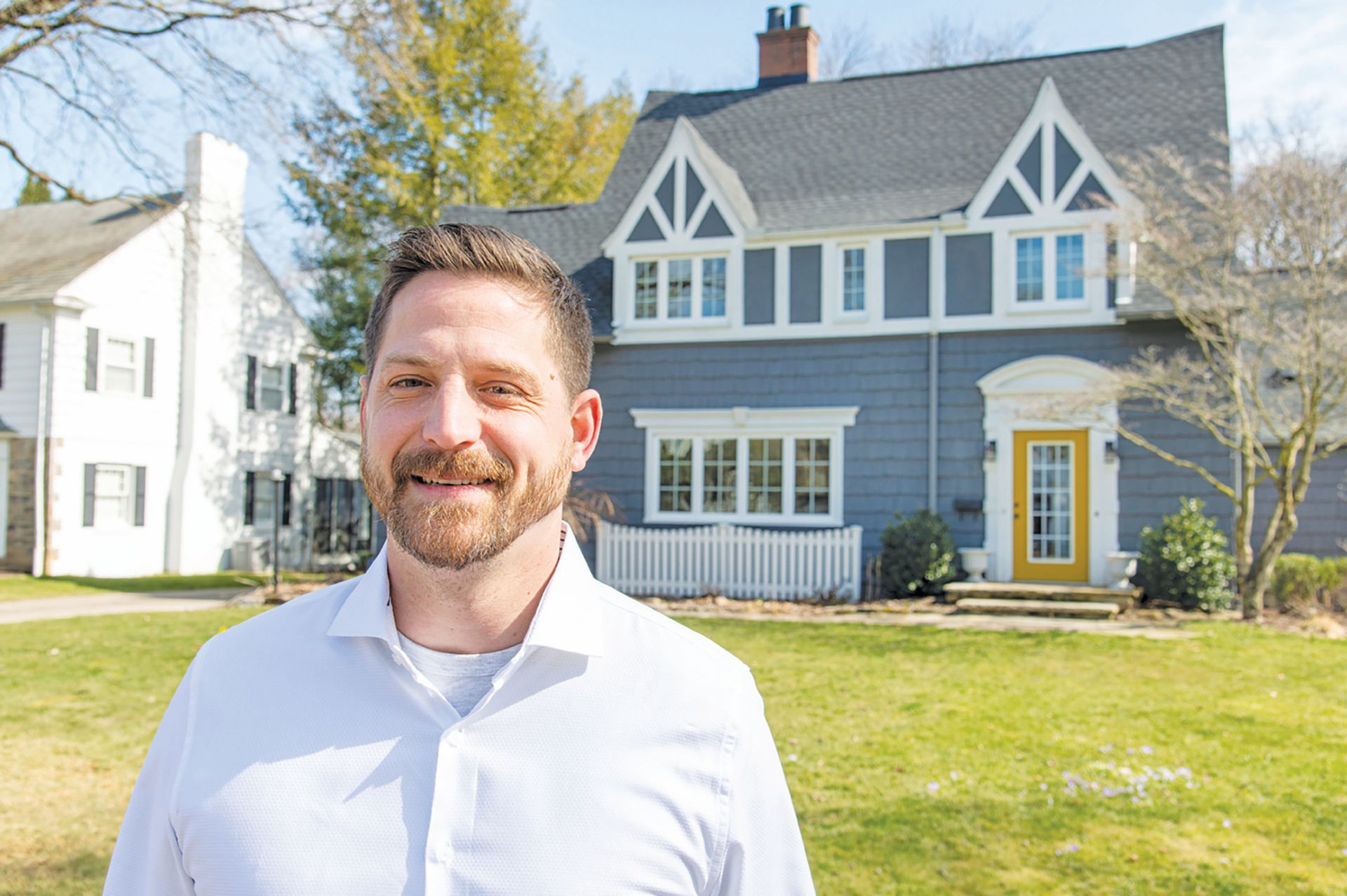 A man smiles in front of a blue two-story house with a yellow front door and a white picket fence on a sunny day.