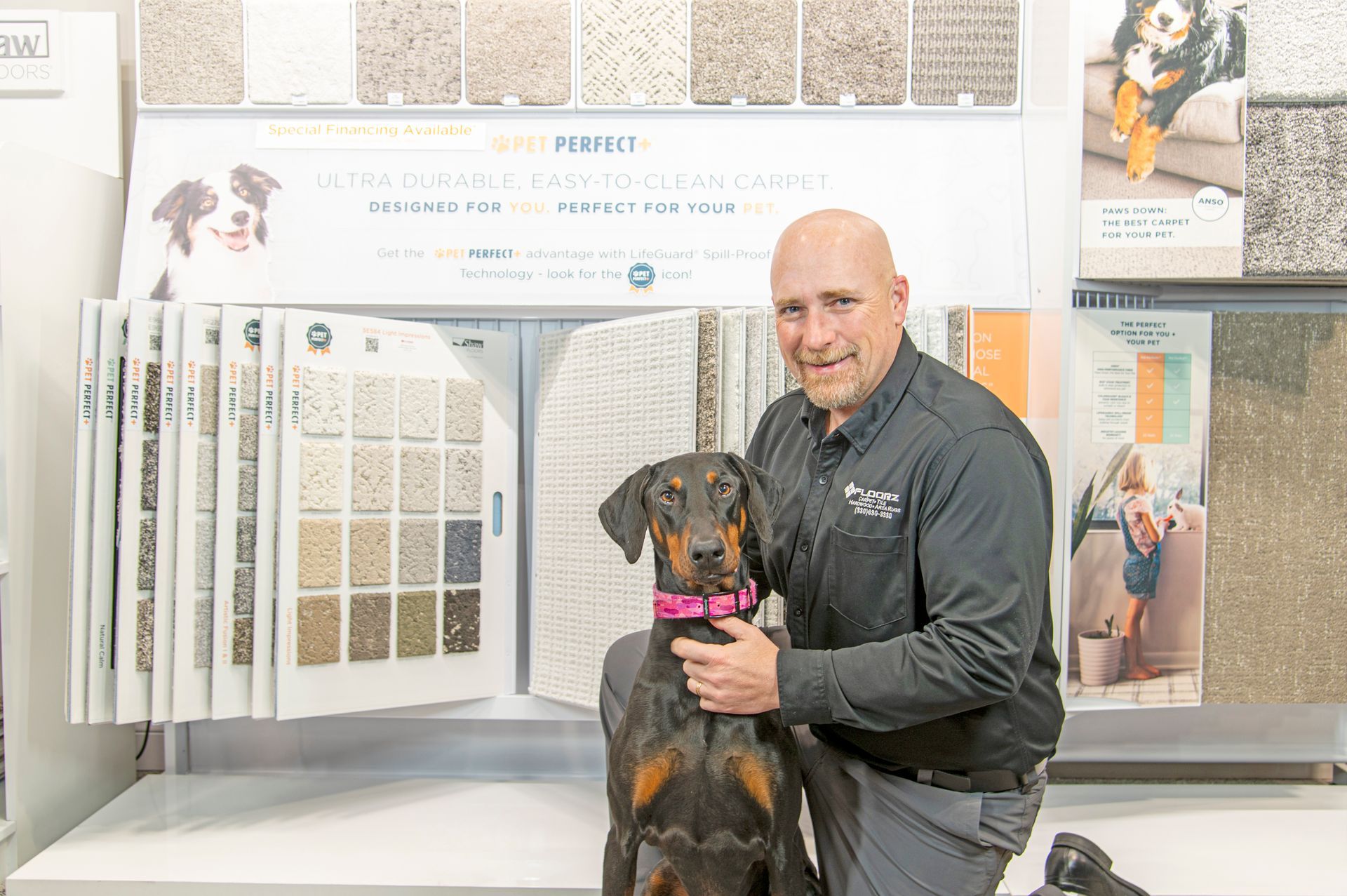 A man with a Doberman Pinscher stands in a carpet showroom in front of displays of various carpet samples.