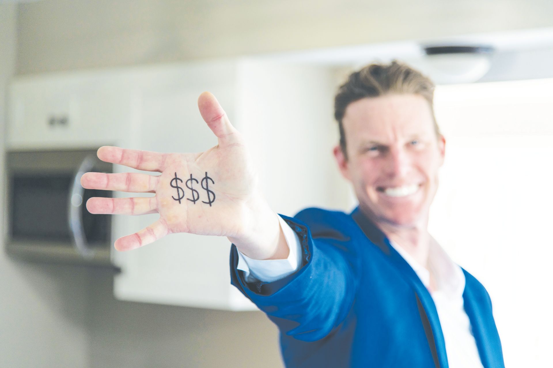 A smiling person in a blue blazer extends their hand toward the camera, which has three dollar signs drawn on the palm.