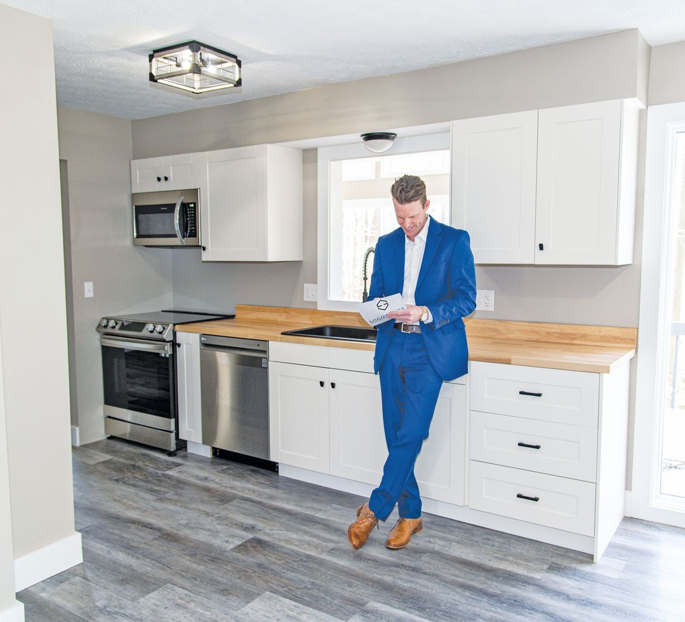 A person in a blue suit leans against a white kitchen counter, looking down at a paper document in a modern kitchen.