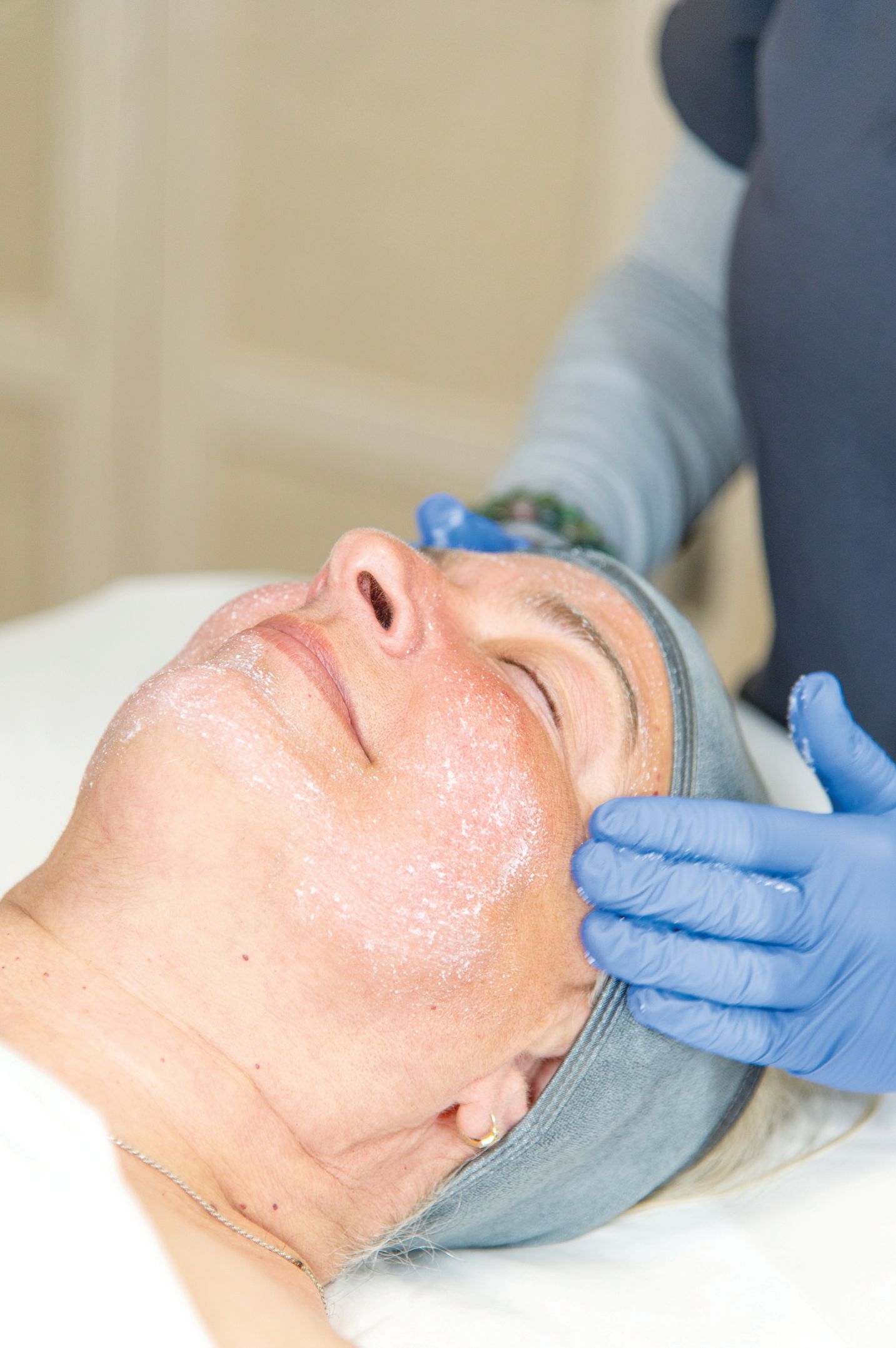 A person in blue gloves performs a facial treatment on a client lying down with a product applied to their face.
