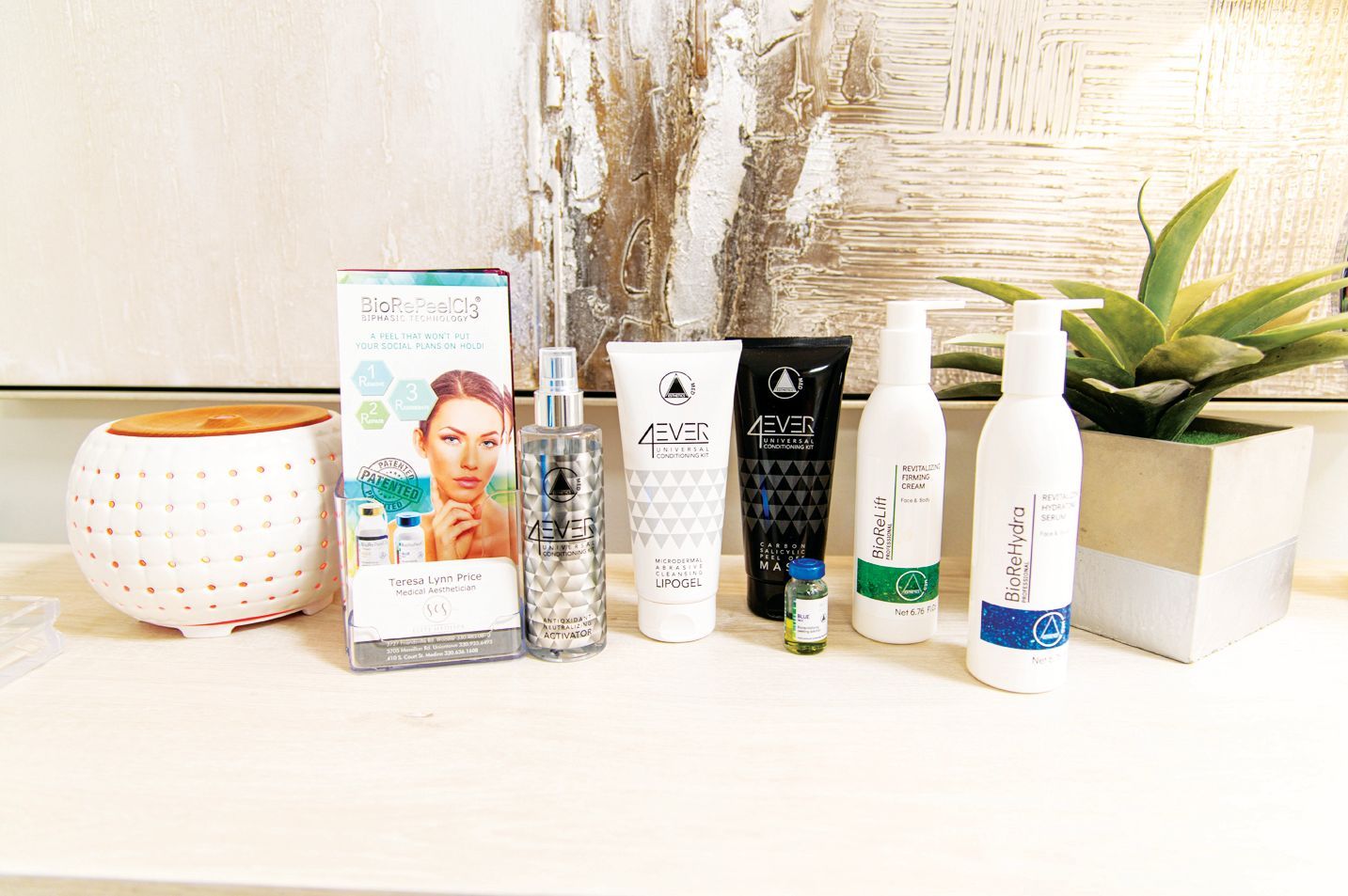A row of various skincare products and a small succulent in a white planter arranged on a white shelf.