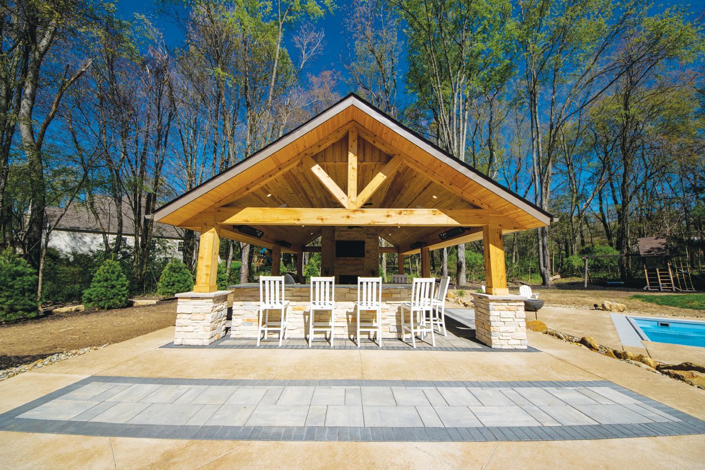 A covered wooden outdoor kitchen and bar structure with four white bar stools, set on a stone patio near a swimming pool.