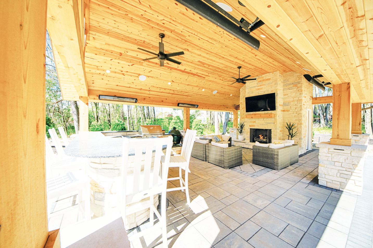 A covered patio with light wood ceilings, fans, a stone fireplace with a mounted TV, and outdoor wicker furniture.