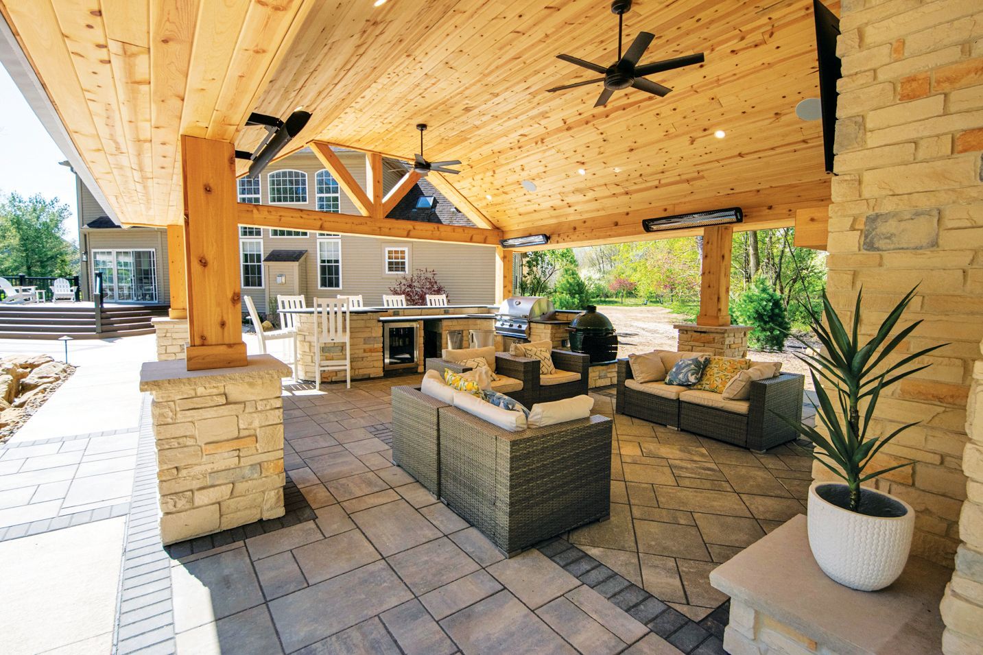 A spacious covered outdoor patio with wood-paneled ceilings, stone pillars, casual seating, and an outdoor kitchen area.