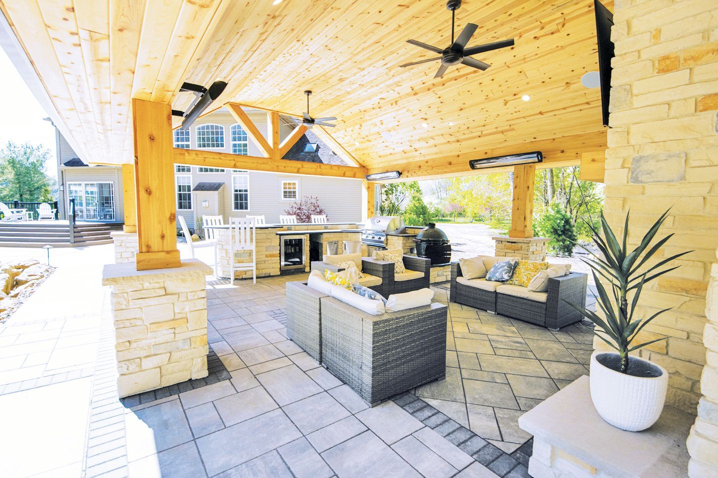 A covered outdoor patio featuring stone pillars, wood ceilings, a lounge area with gray furniture, and a built-in kitchen.