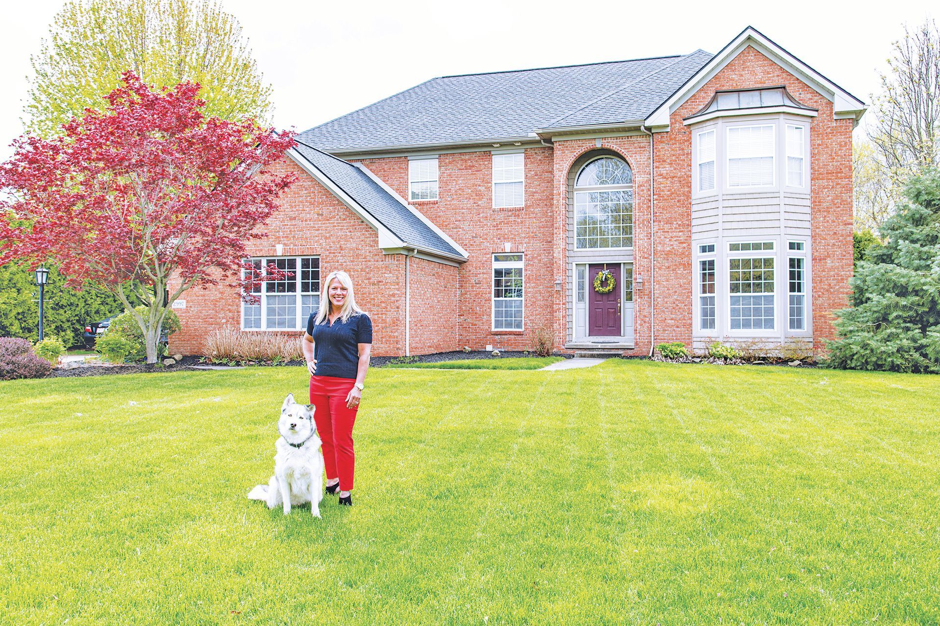 A person stands on a lush lawn in front of a two-story brick house with a white dog sitting beside them.