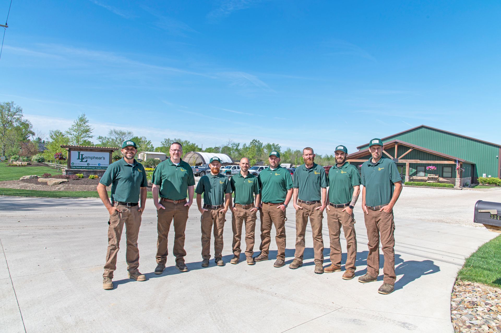Nine people in green shirts and khaki pants standing in a line outdoors in front of a building on a sunny day.