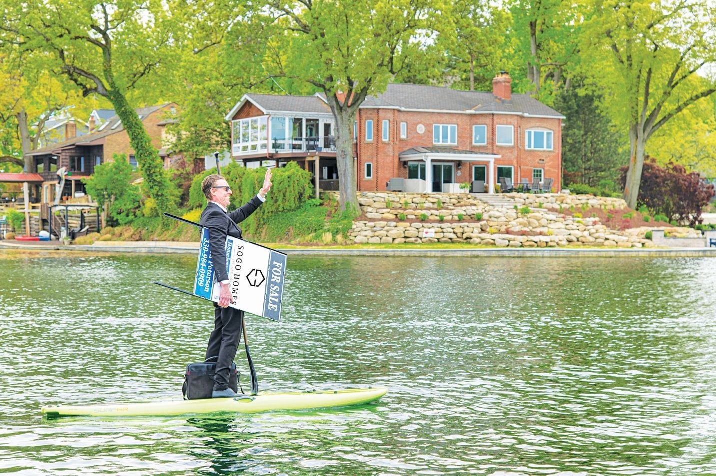 A person in a suit stands on a paddleboard on a lake, waving, while holding a sign with a logo, in front of a brick house.