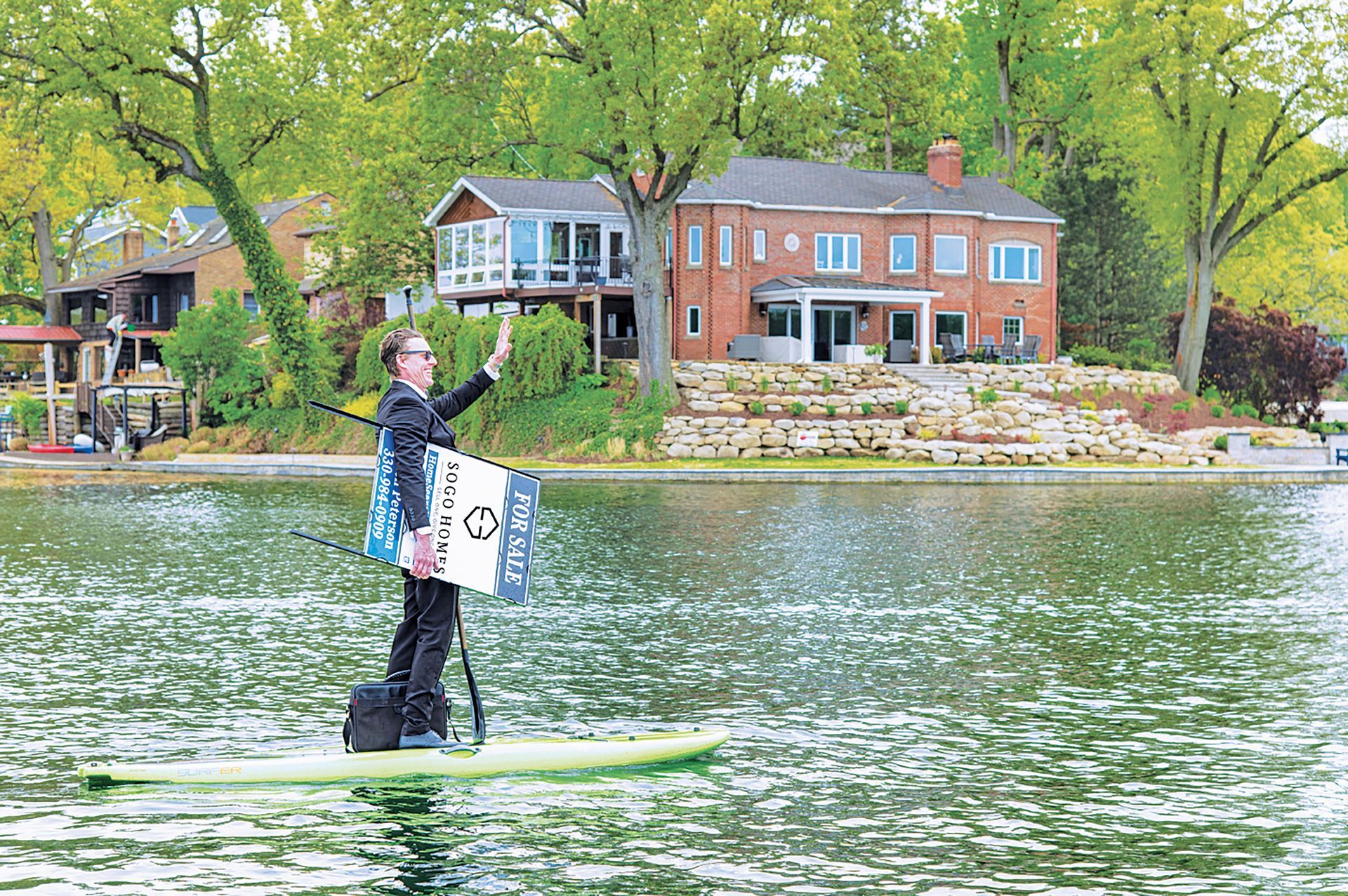 A person in a suit stands on a paddleboard on a lake, waving, while holding a campaign sign in front of a house.