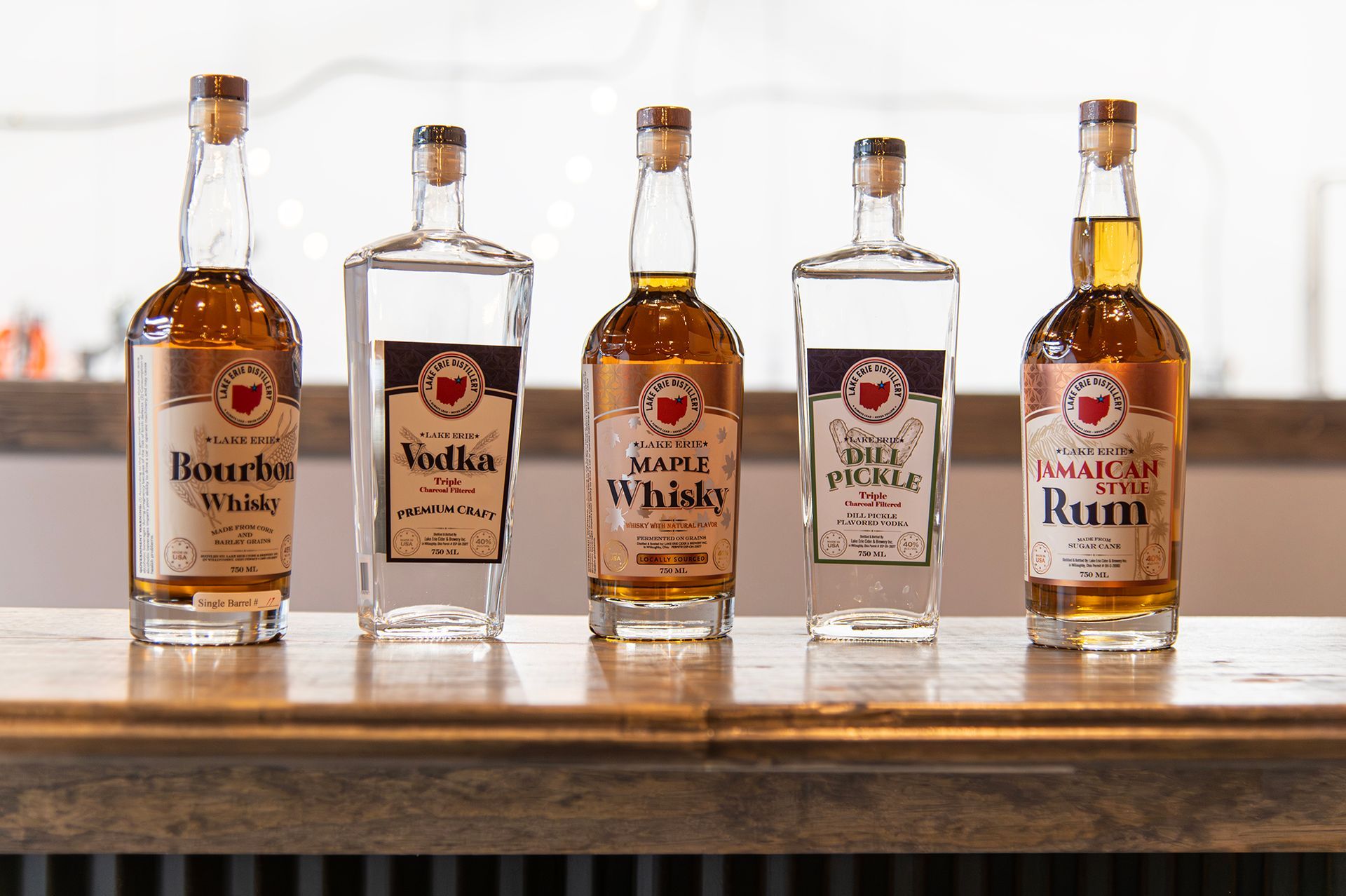 Five bottles of craft spirits with labels featuring the state of Ohio, lined up on a wooden bar top.