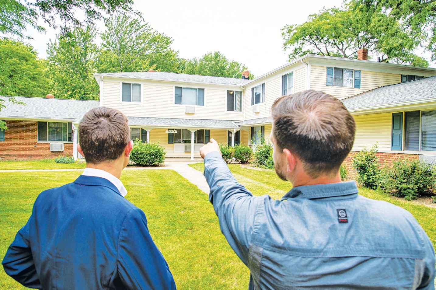 Two people stand on a grassy lawn looking at a multi-story house, with one person pointing toward the building.