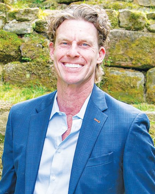 A smiling person with wavy hair wearing a blue blazer and light blue button-down shirt, standing before a stone wall.