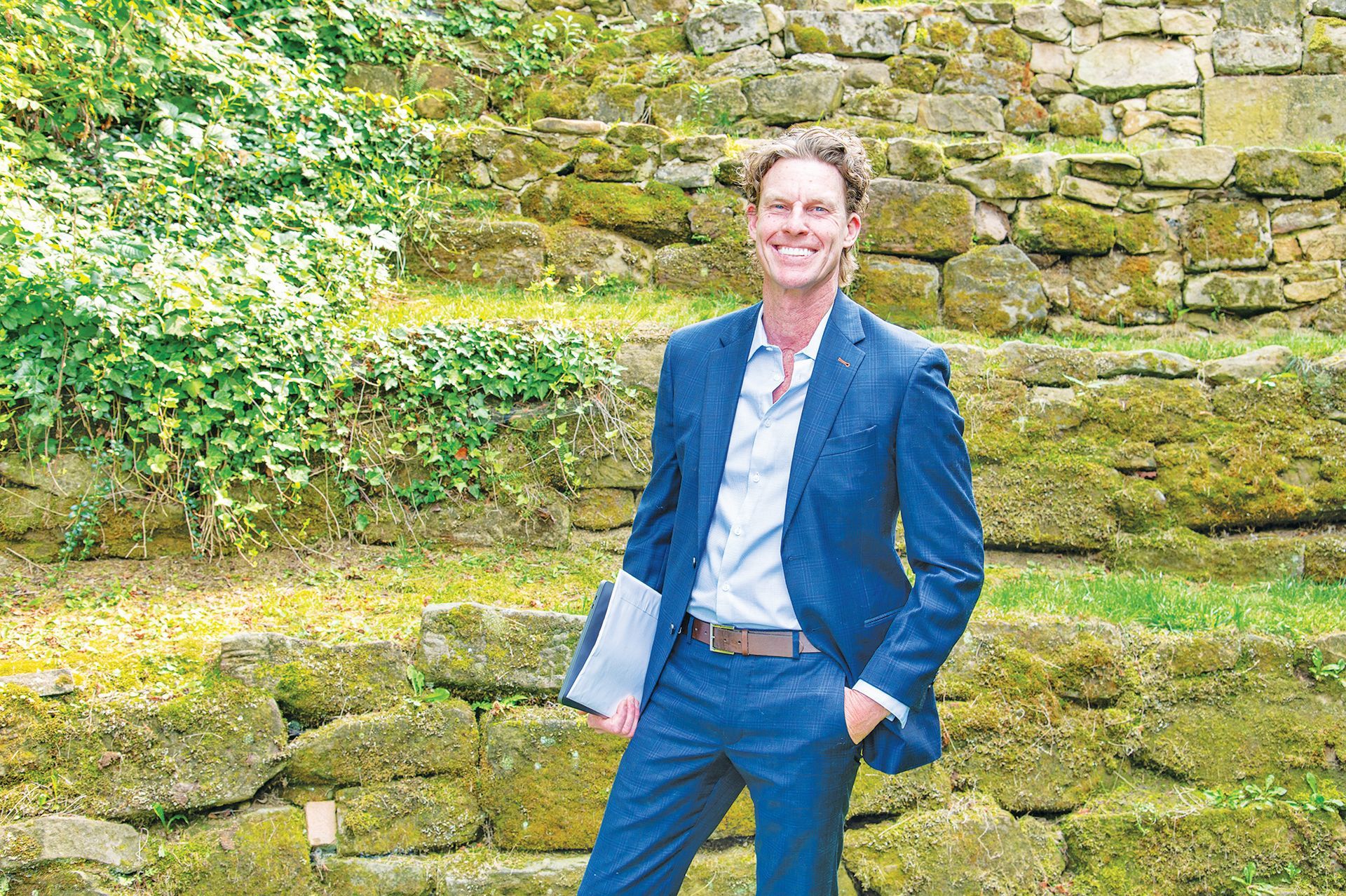 A smiling person in a blue suit stands in front of a terraced stone wall covered in moss and lush greenery.