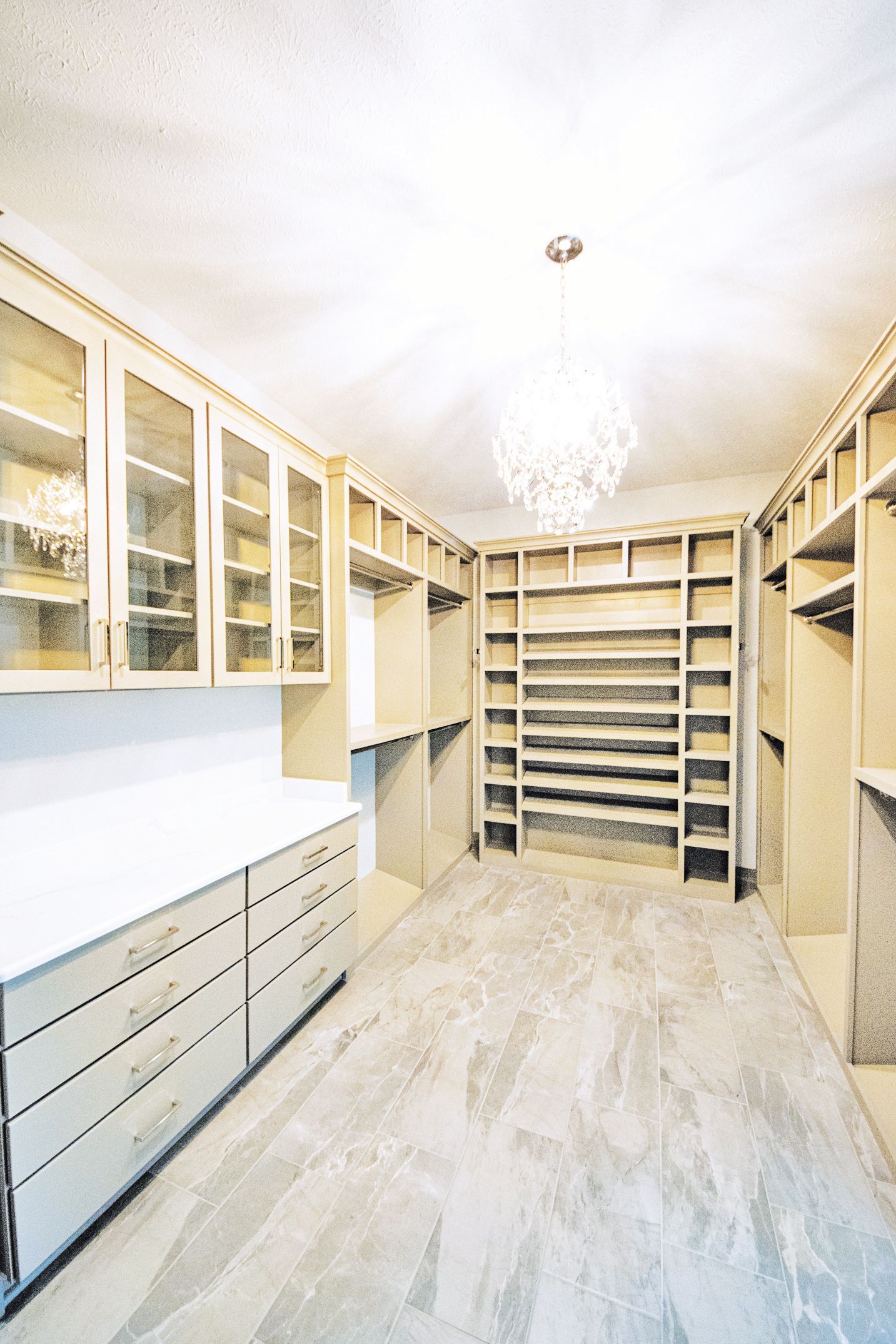 A walk-in closet featuring light wood shelving, glass-front cabinets, white drawers, and a crystal chandelier.