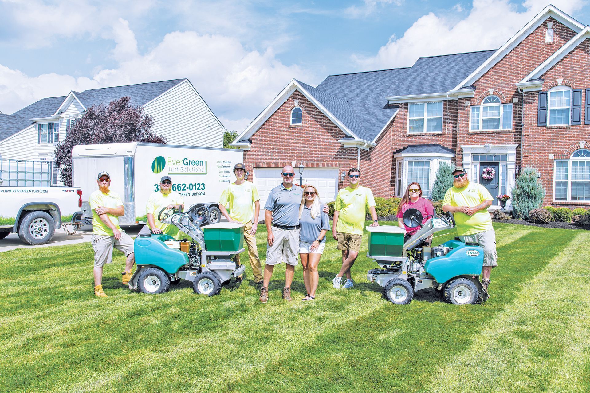 A group of nine professionals in bright yellow and casual attire stand in a residential lawn with lawn care equipment.