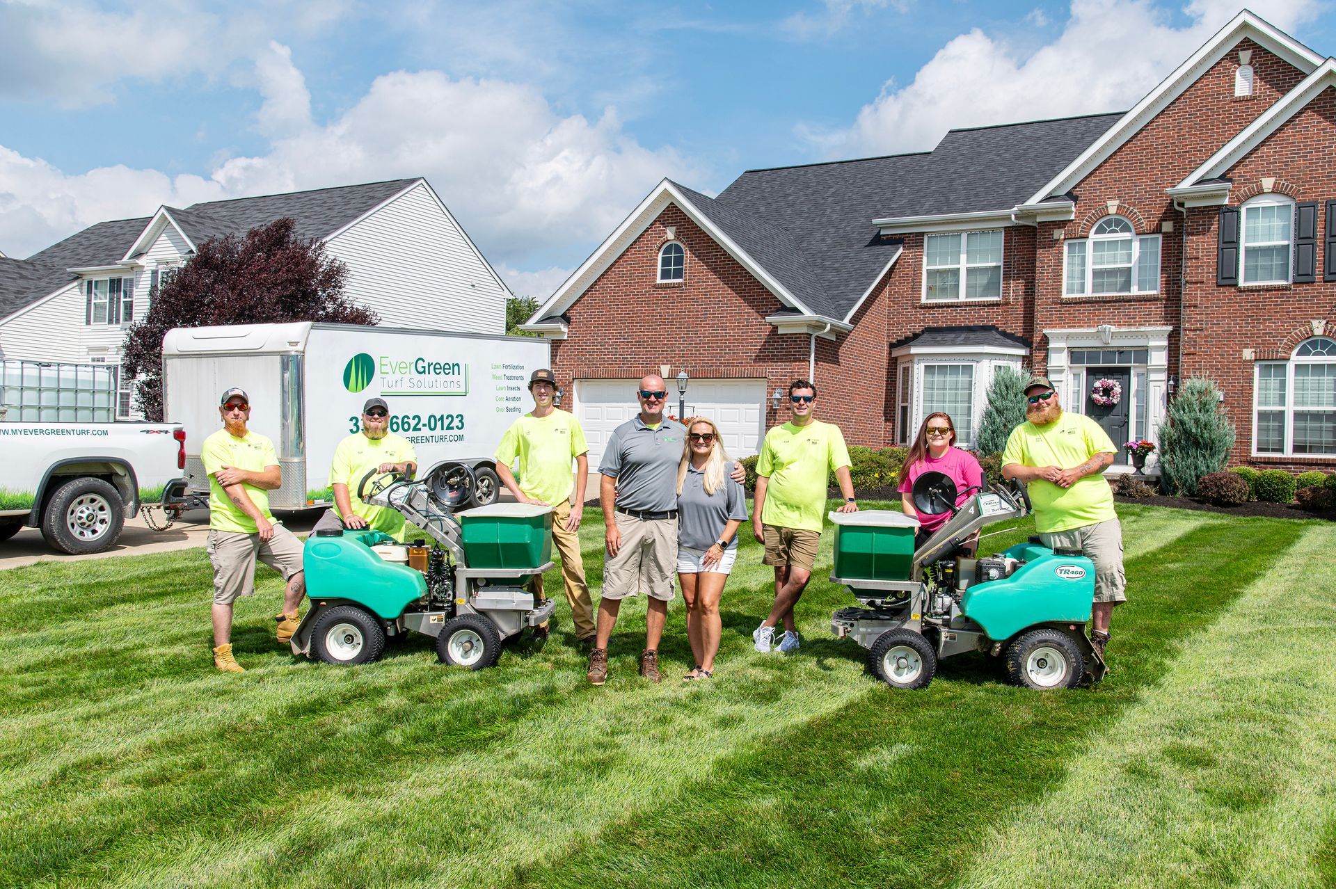 A team in neon yellow and casual shirts posing with turf equipment on a green lawn in front of a brick house.
