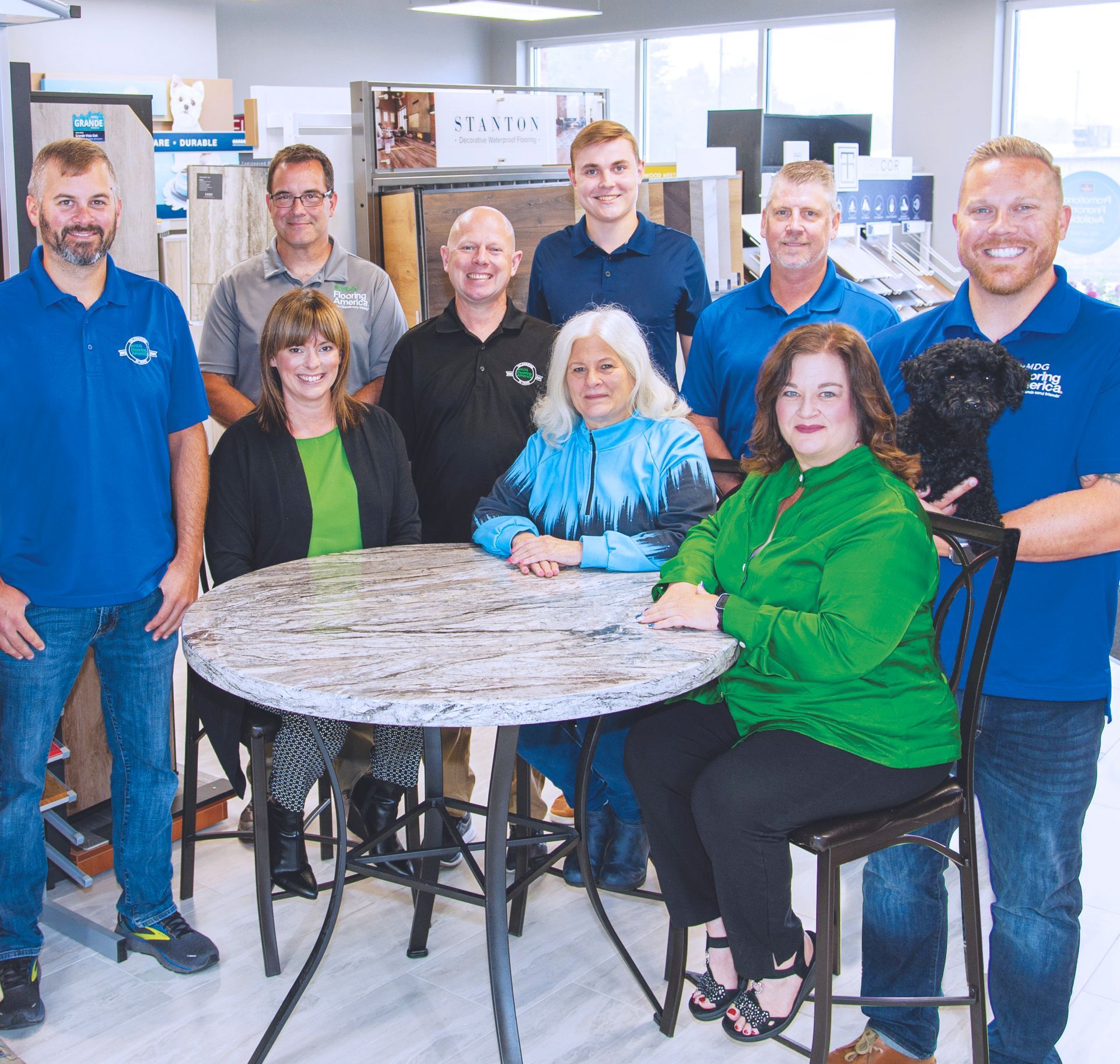 A group of ten people and a small dog posing for a photo in a showroom with a round, stone-top table in the center.