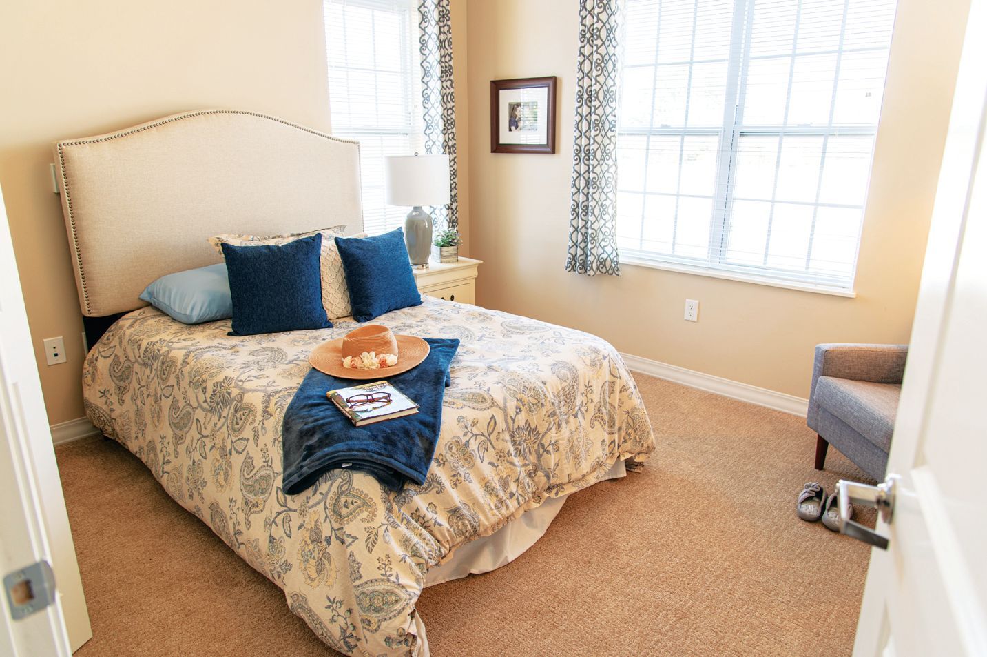 A beige bedroom with a bed featuring a cream headboard, patterned quilt, blue pillows, and a straw hat on a blue throw.