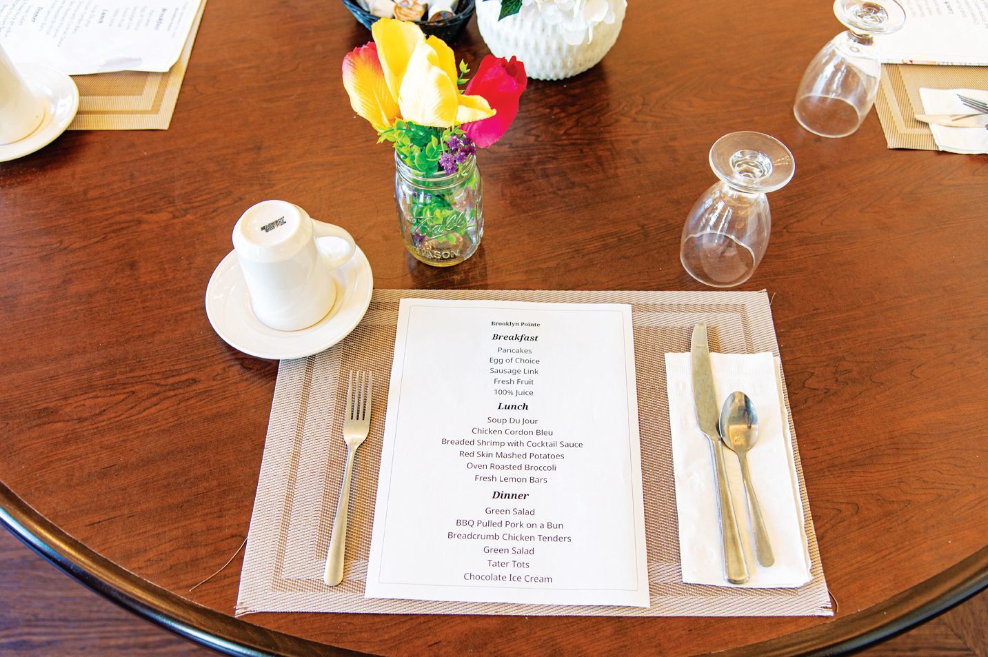 A dining table setting with a menu, silverware, a coffee cup, and a small glass vase of colorful flowers.