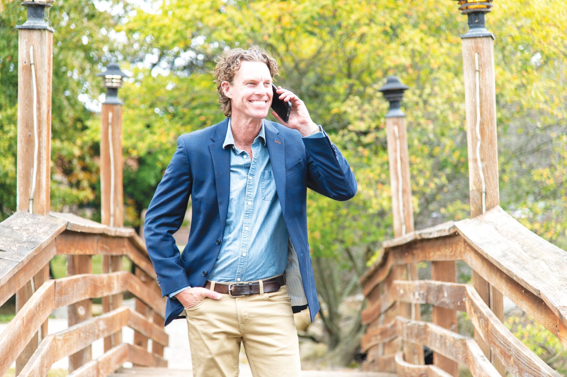 A person in a blue blazer and tan pants walks across a wooden bridge while talking on a smartphone.
